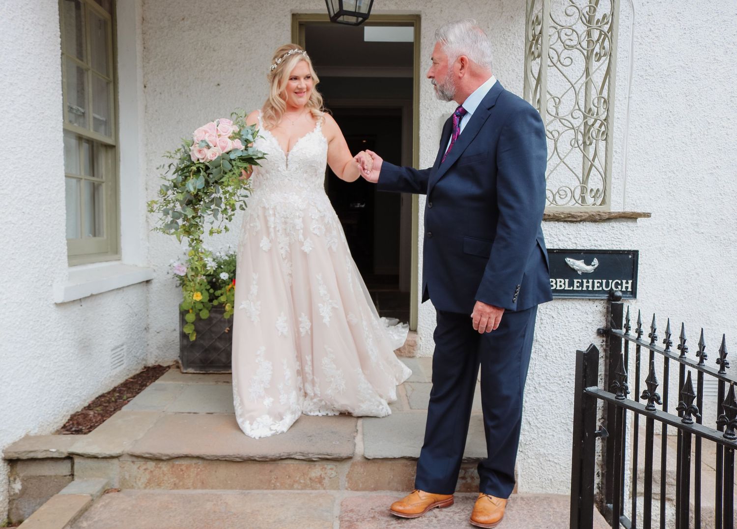 Two people share a touching moment on a white porch, with one wearing a flowing lace wedding dress and another in a navy suit.