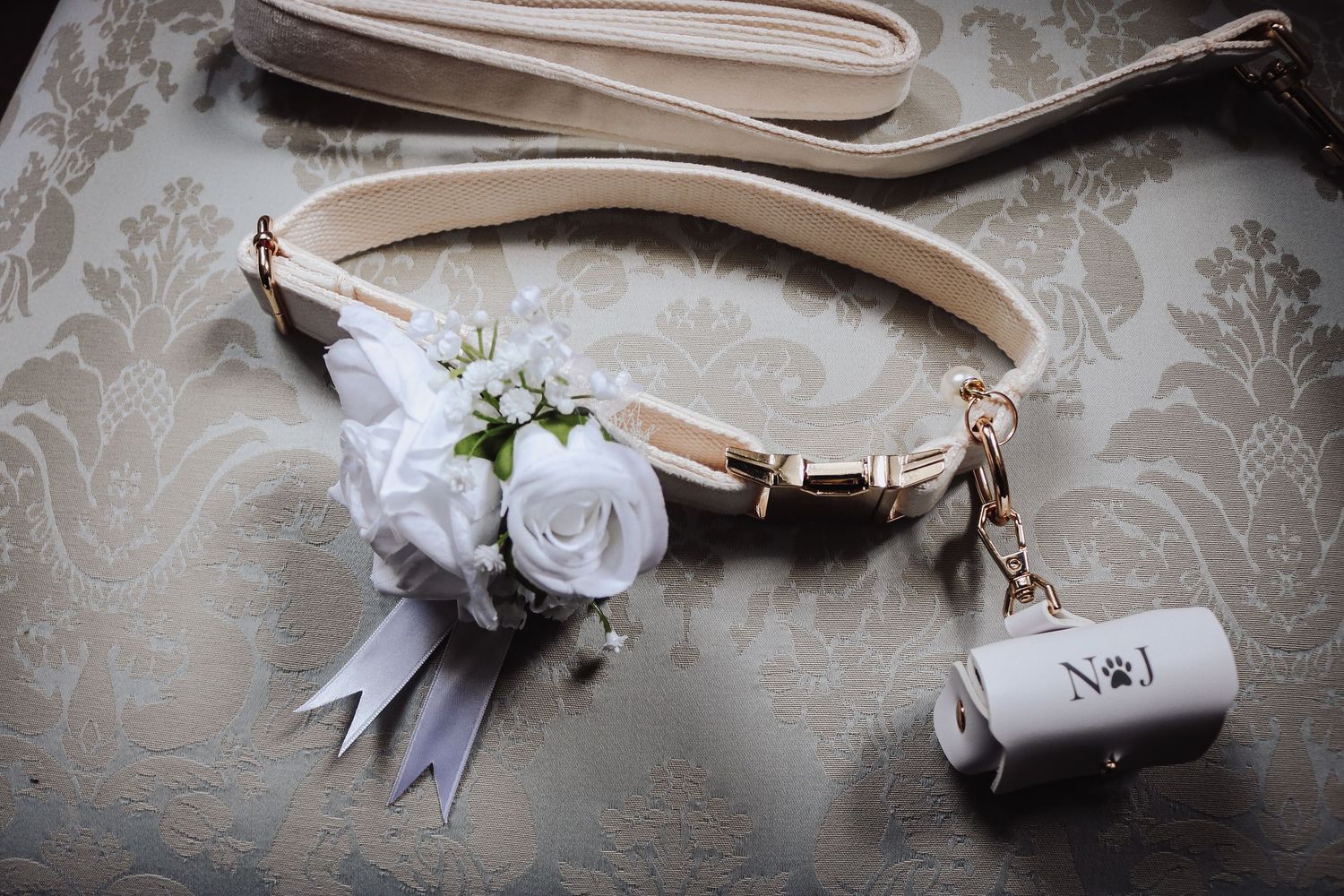 Elegant pet collar decorated with white roses and ribbons on a beige damask background with a small bag charm attached.