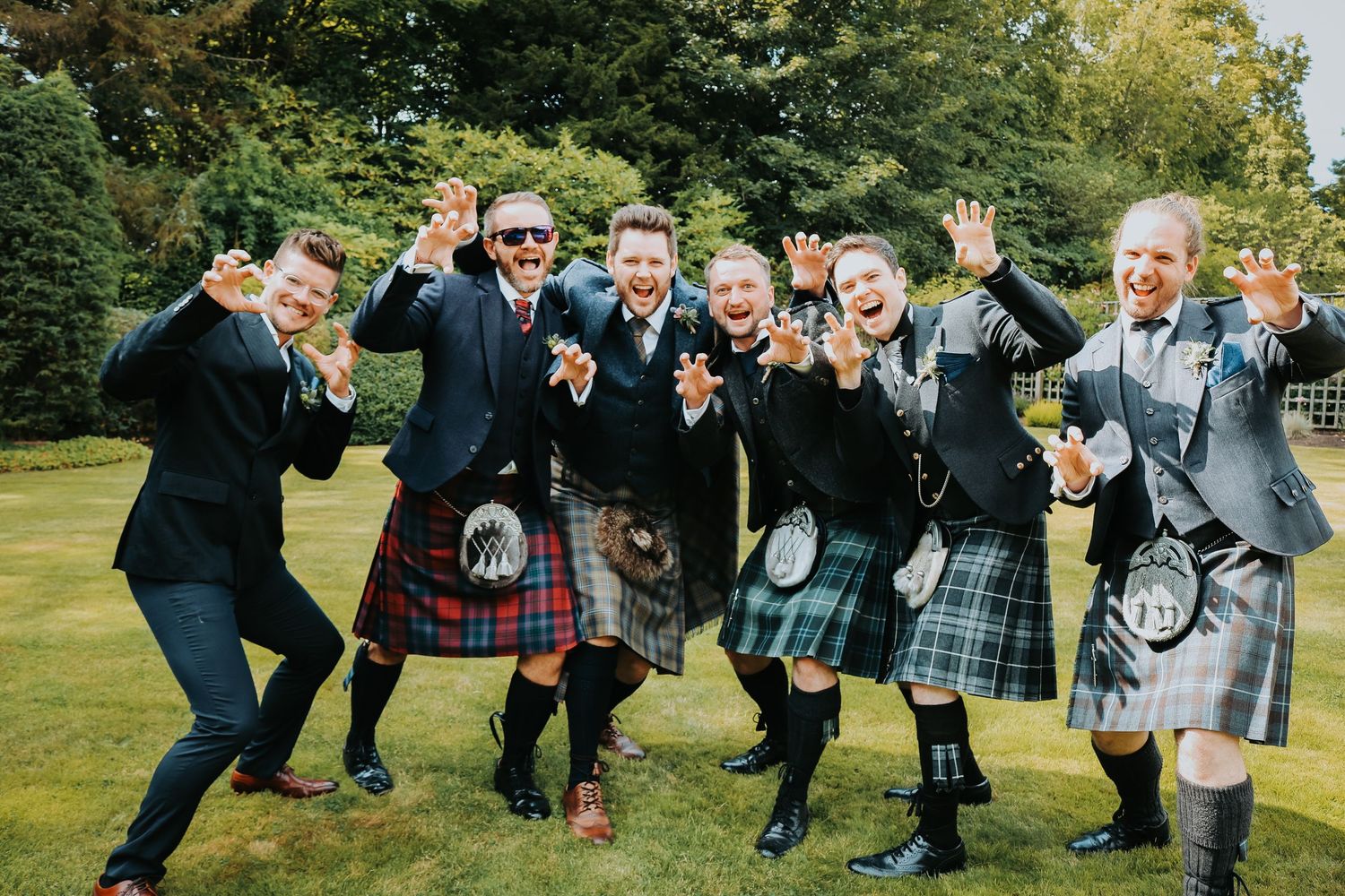 A cheerful group in traditional Scottish kilts strike an energetic pose together on a lawn surrounded by trees.