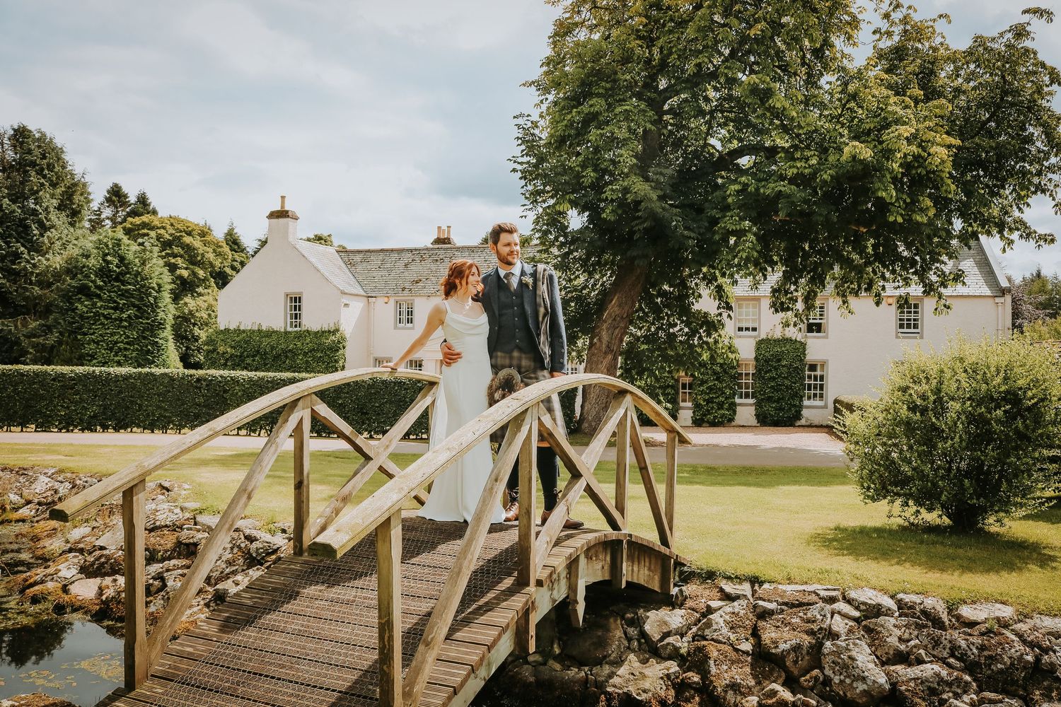 A couple in wedding attire stands on a charming wooden footbridge in a lush garden setting on their special day.