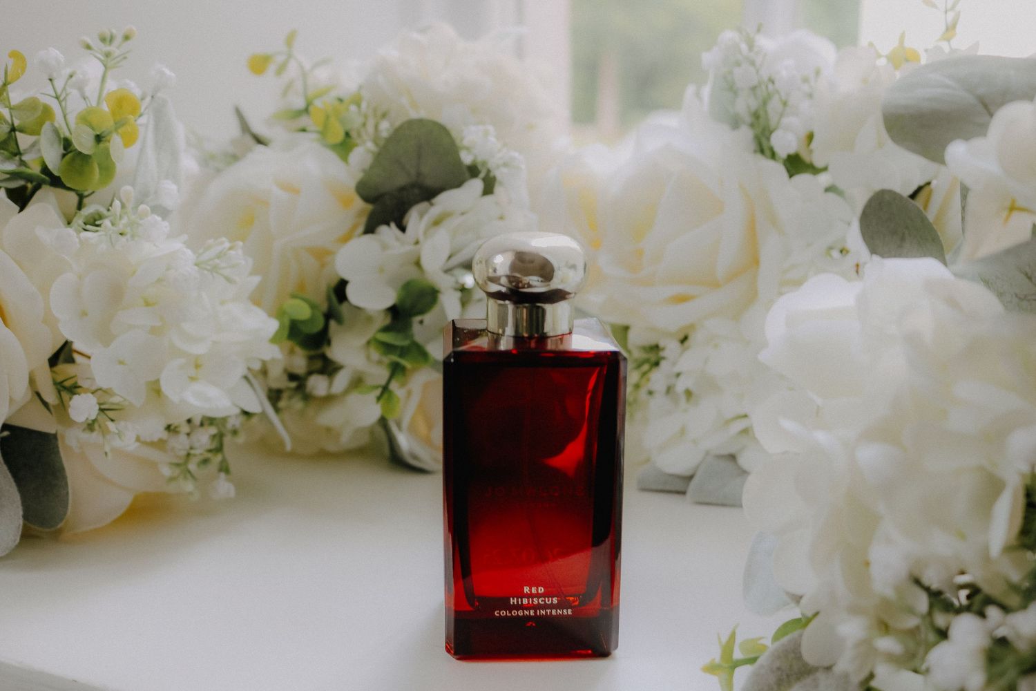 A luxury perfume bottle with red liquid sits among delicate white flowers and greenery in an elegant display.