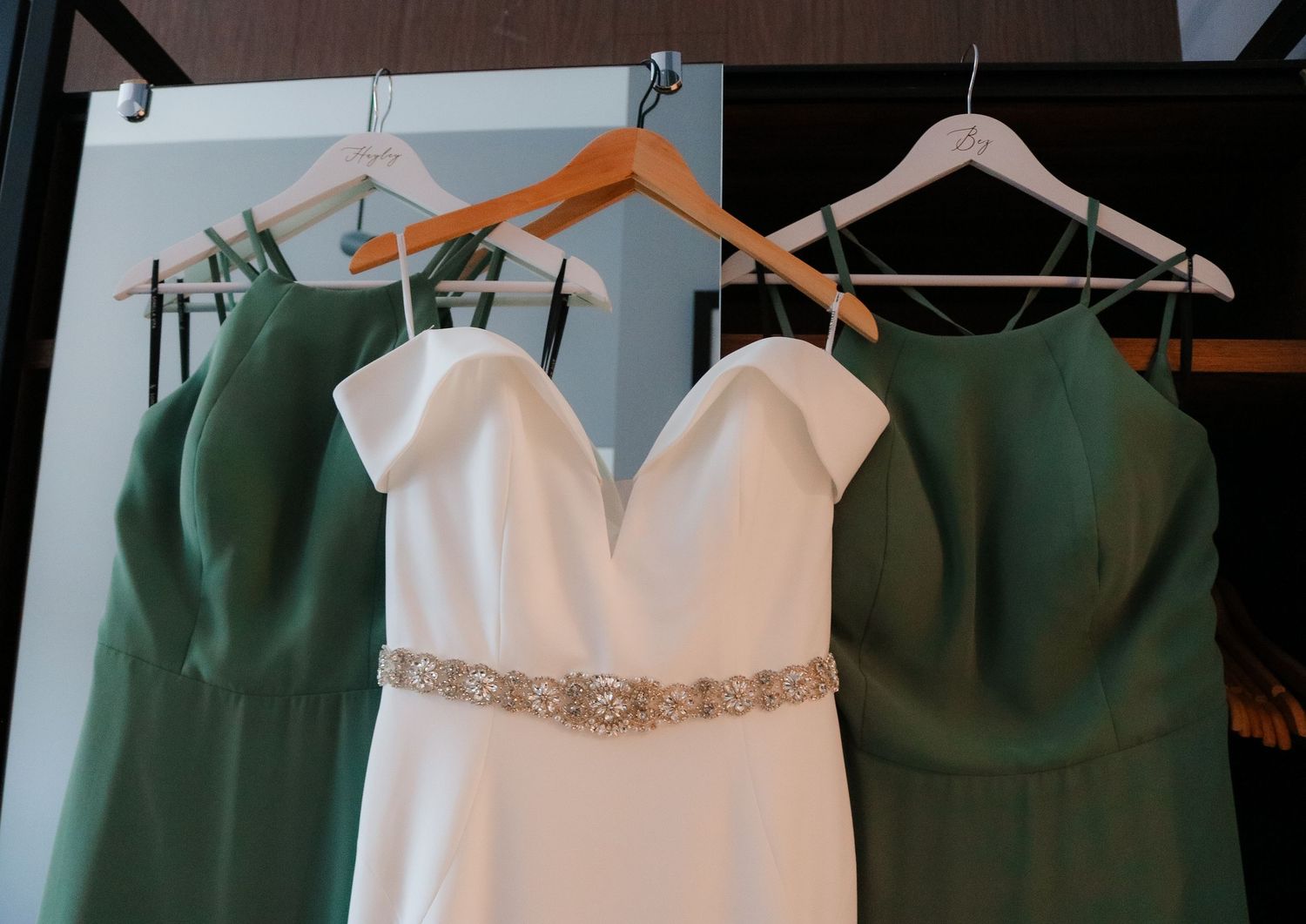 Three formal dresses hanging on wooden hangers display emerald green and white colors with delicate beaded details.
