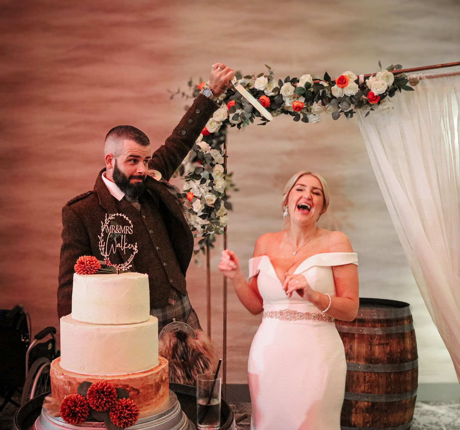 A joyful wedding celebration moment at the reception with a white tiered cake decorated with red roses near wine barrels.