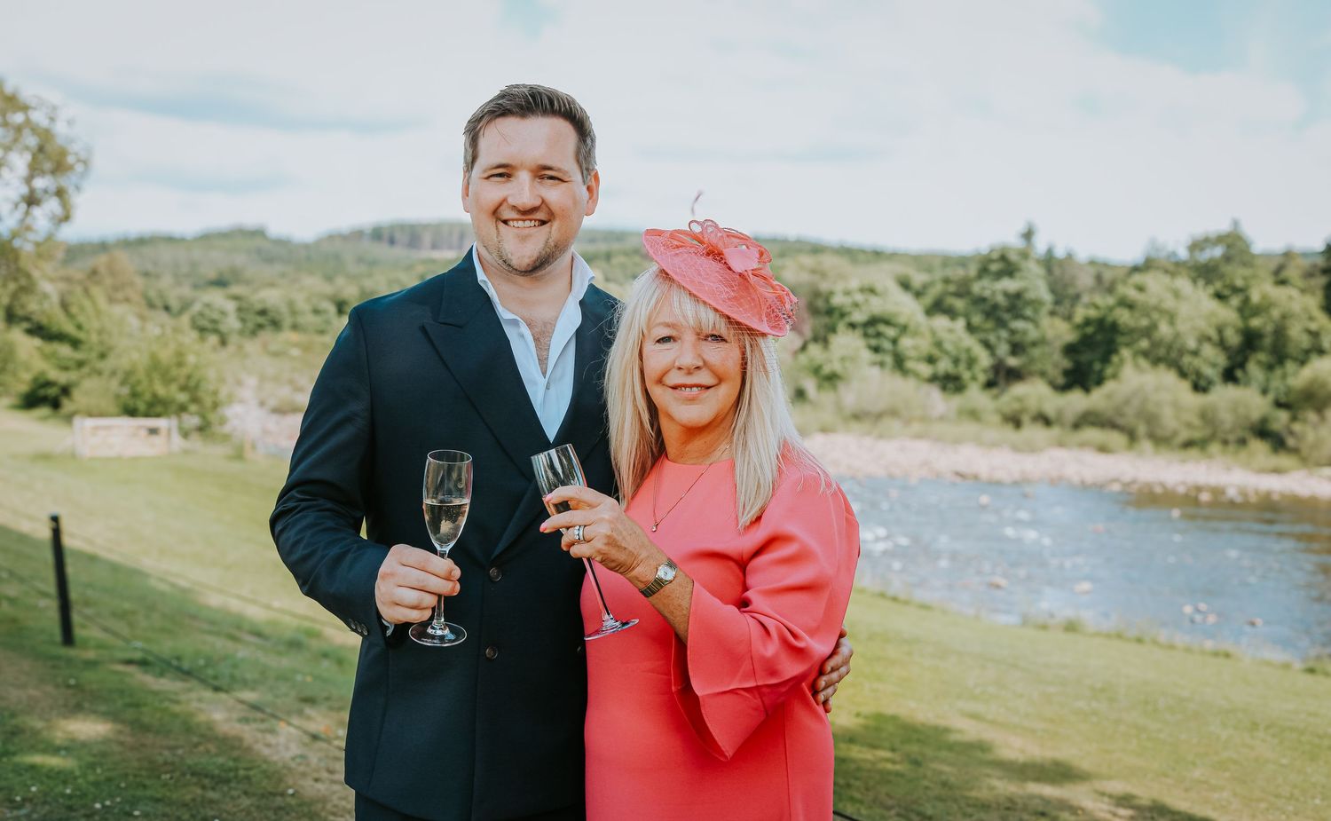 Two people in formal attire hold champagne glasses while celebrating at an outdoor venue near a river on a sunny day.