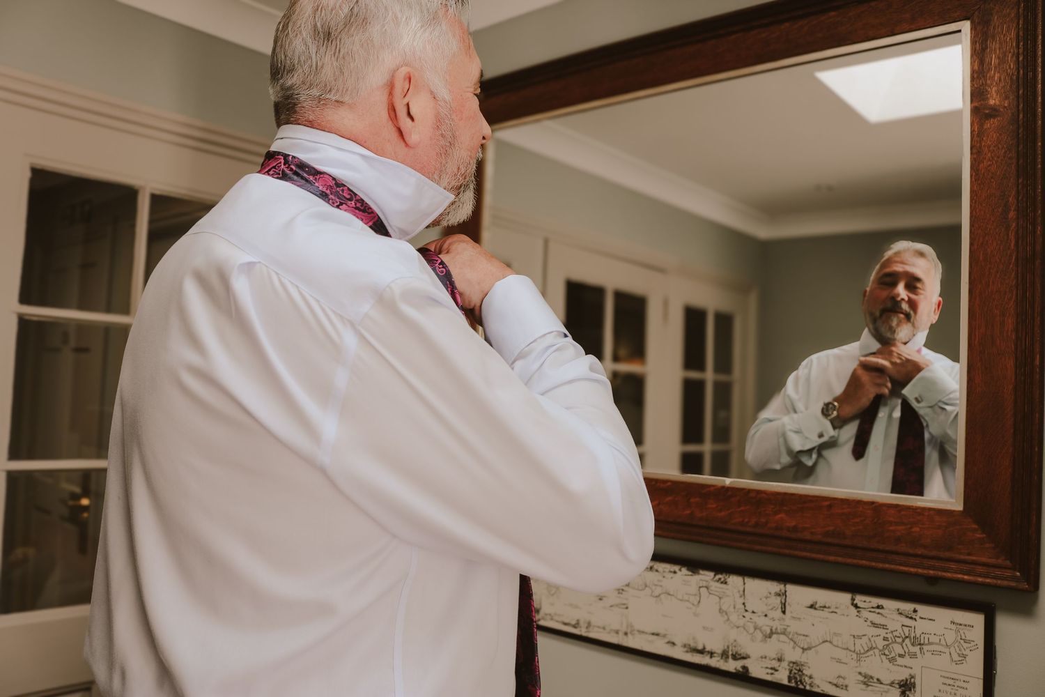 Someone in a white dress shirt and tie adjusts their collar while looking into a bathroom mirror.
