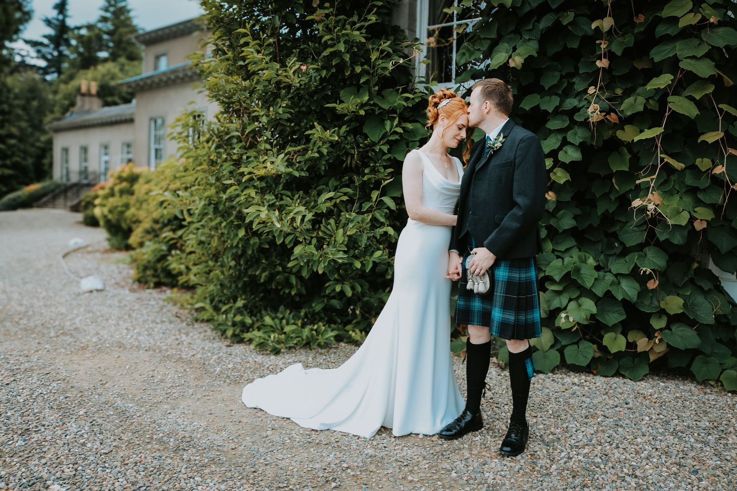 A couple shares a romantic moment in front of green foliage, with the groom wearing a traditional Scottish kilt.