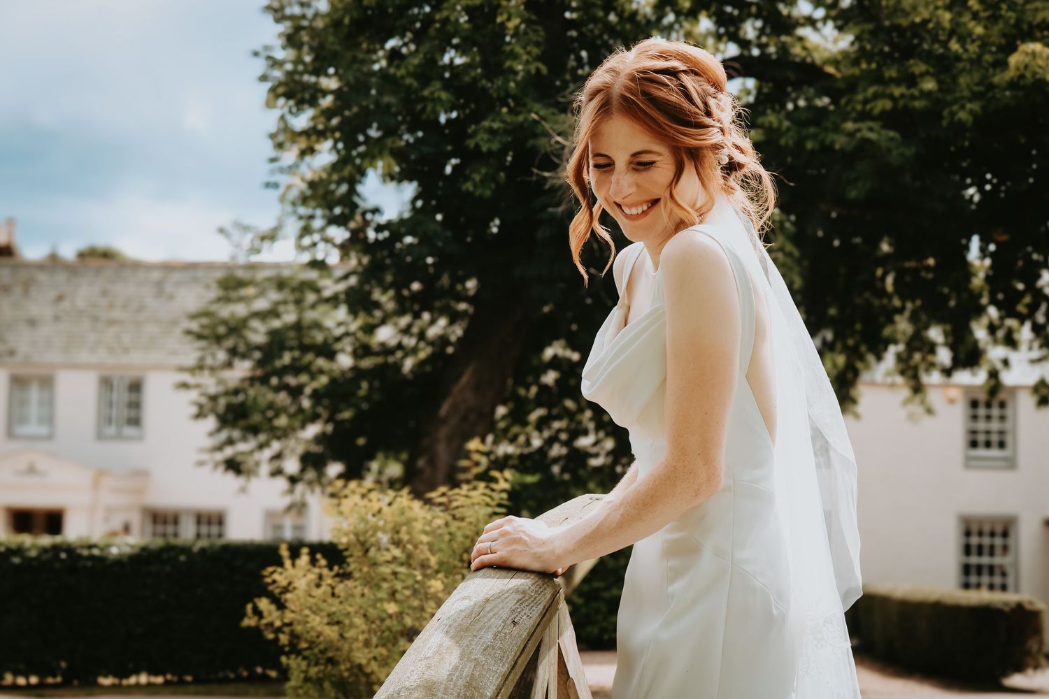 A bride in an elegant white wedding dress stands in a sunlit garden setting with classic architecture in the background.