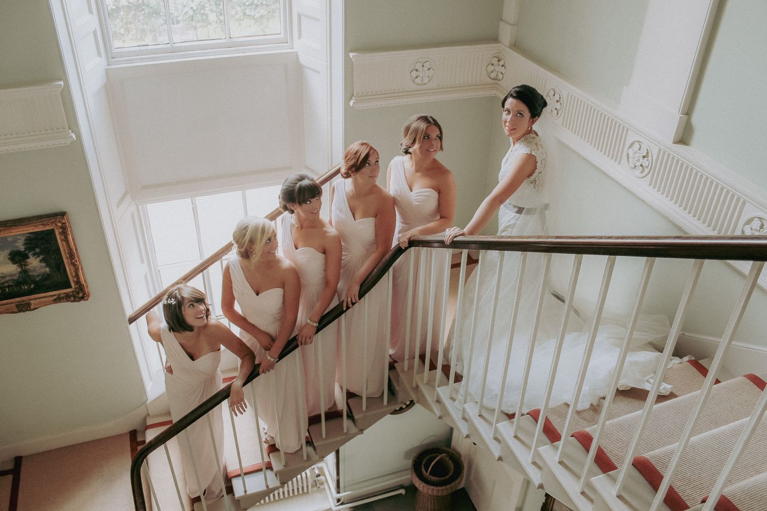 A group in blush pink dresses stands elegantly arranged on a white staircase in a classic indoor setting.
