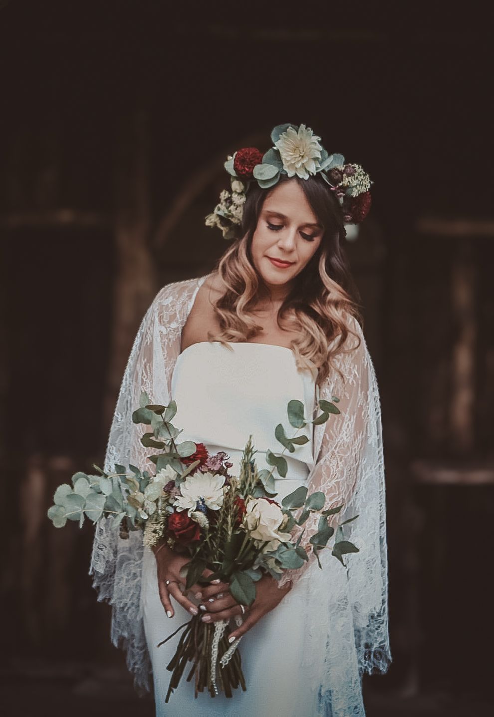 Bride in white lace dress and floral crown holds elegant burgundy and eucalyptus bouquet in moody dark setting.