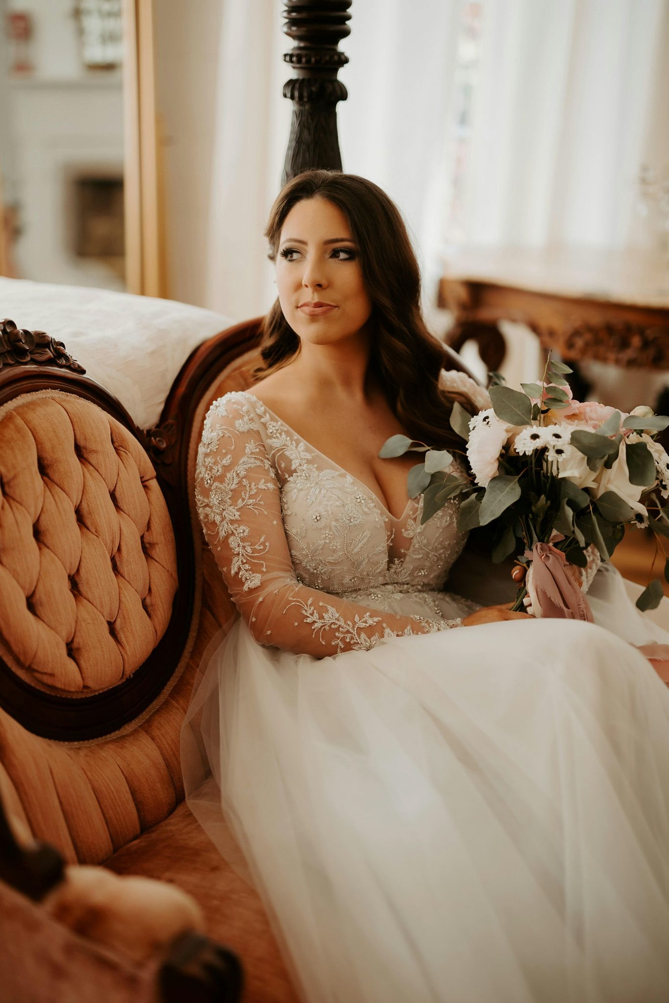 A bride in a long-sleeved lace wedding gown sits elegantly on a vintage tufted chair holding a white floral bouquet.