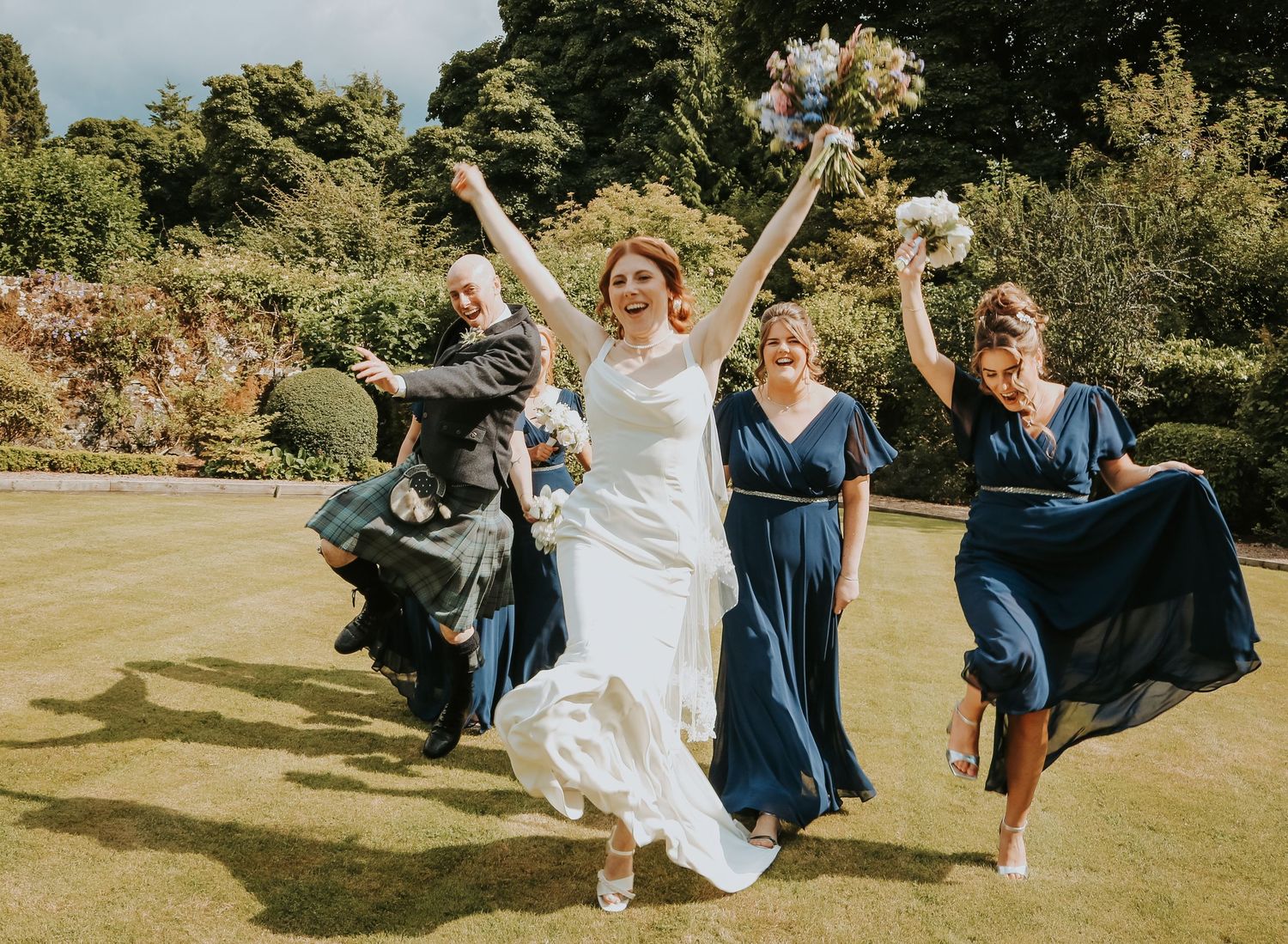 Wedding party in white and navy blue dresses joyfully leaping together on a garden lawn during celebration.
