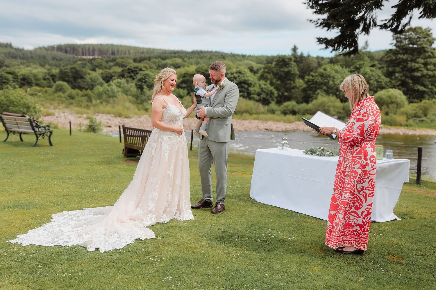 A wedding party gathers on a scenic grassy hilltop overlooking a coastal landscape on a sunny summer day.