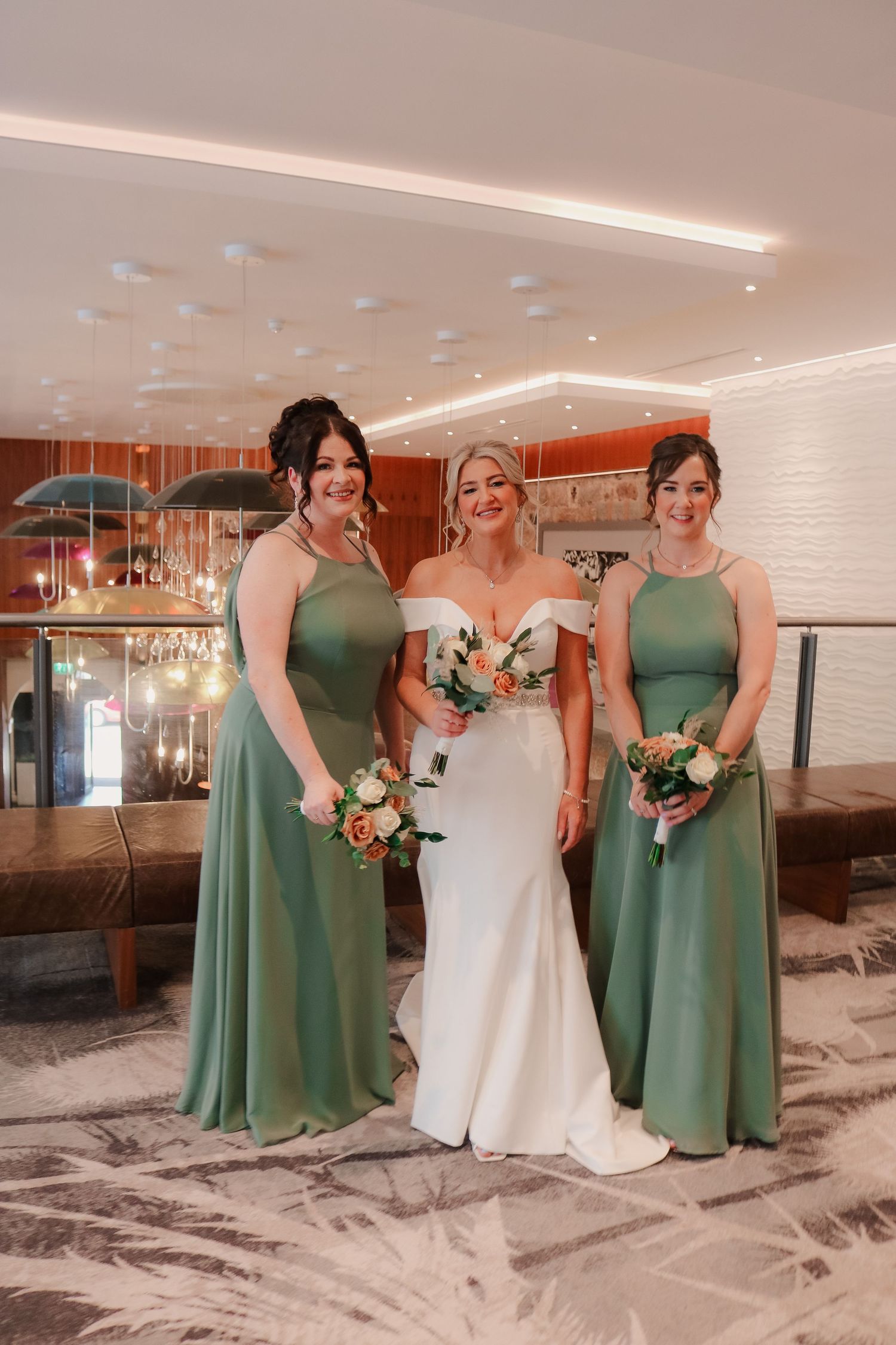 A bridal party poses in a modern venue, with bridesmaids in sage green dresses and the bride in a fitted white gown.