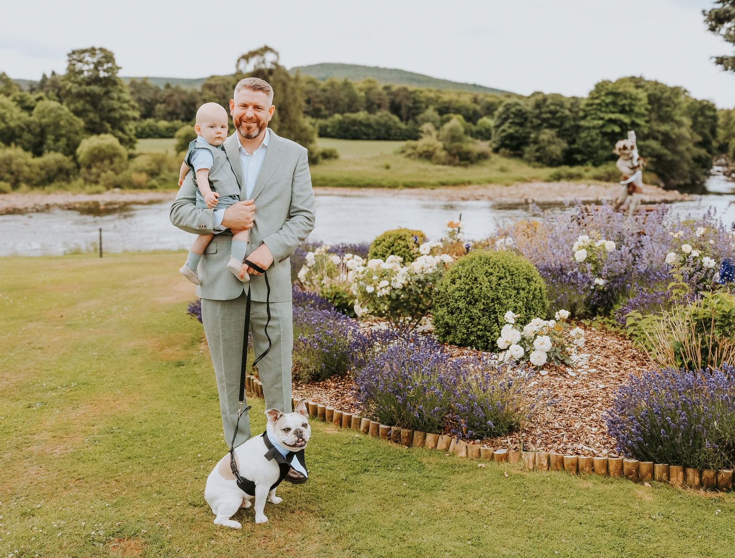 A scenic garden by a lake features blooming lavender flowers and a small dog on a leash against a rolling landscape backdrop.