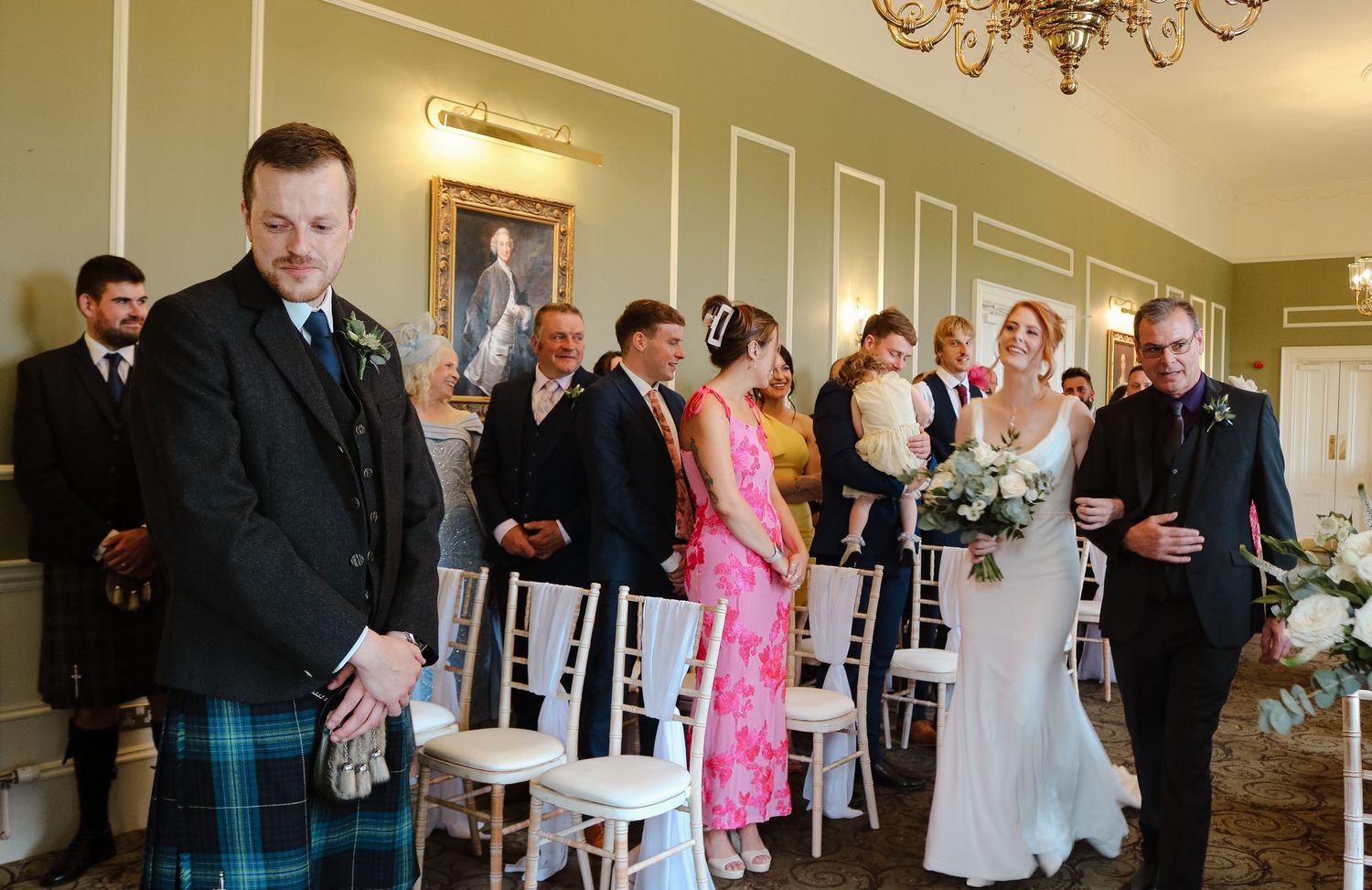 Wedding ceremony taking place in an elegant room with white chairs and a chandelier, featuring Scottish formal wear.