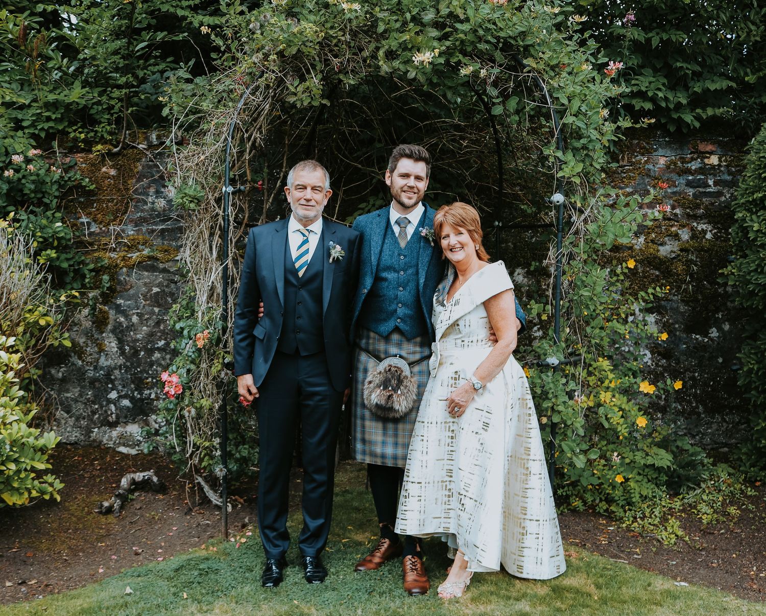 Wedding party poses under a floral archway in a garden setting, with Scottish tartan and elegant attire.