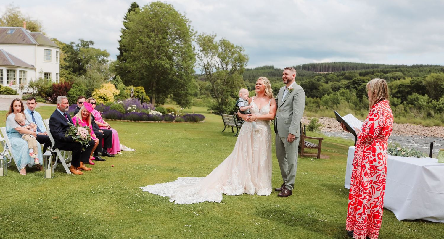 An outdoor wedding ceremony takes place on a lush green lawn with rolling hills in the background on a sunny day.