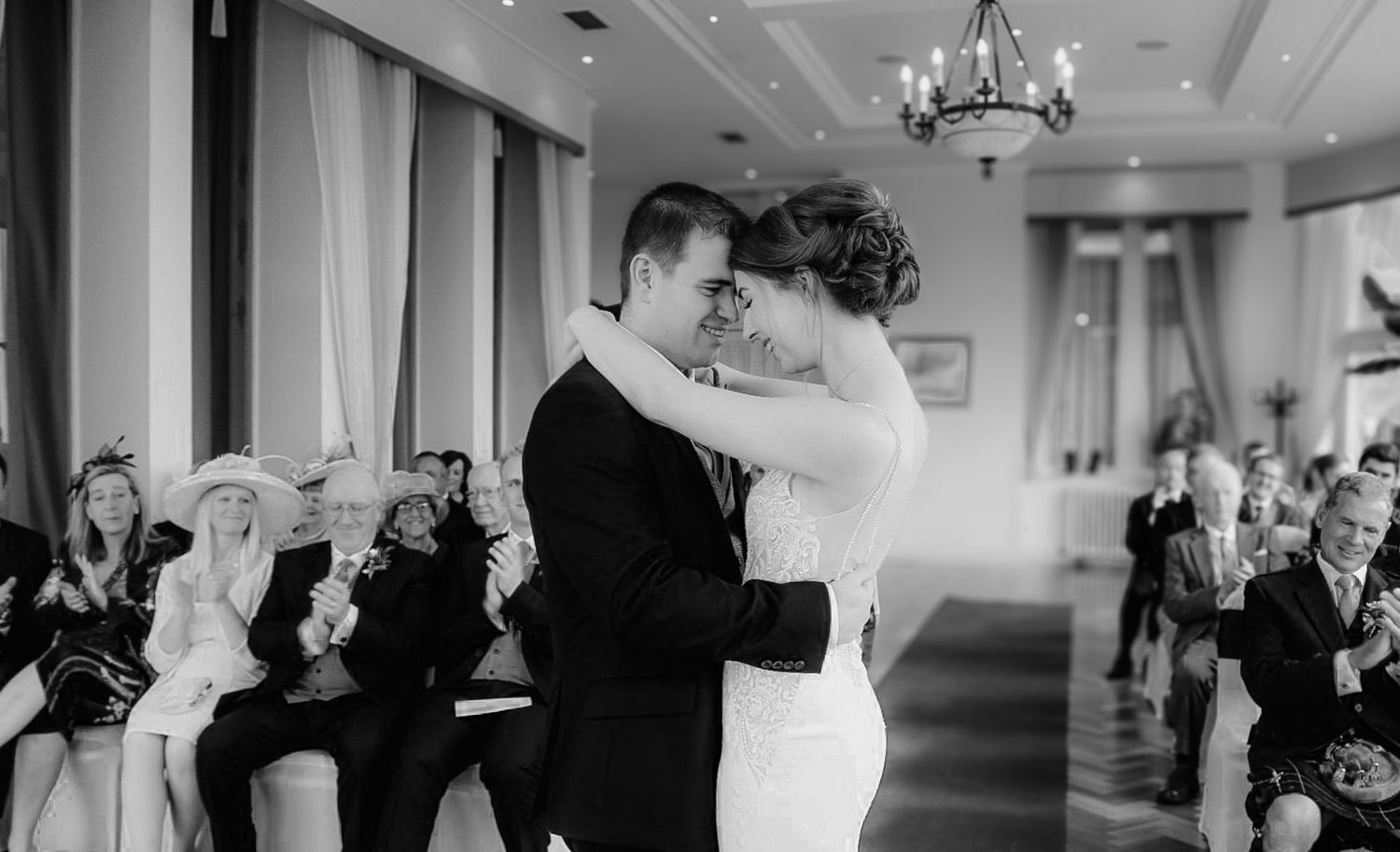 A bride and groom share their first dance at a wedding reception while guests look on in black and white photography.