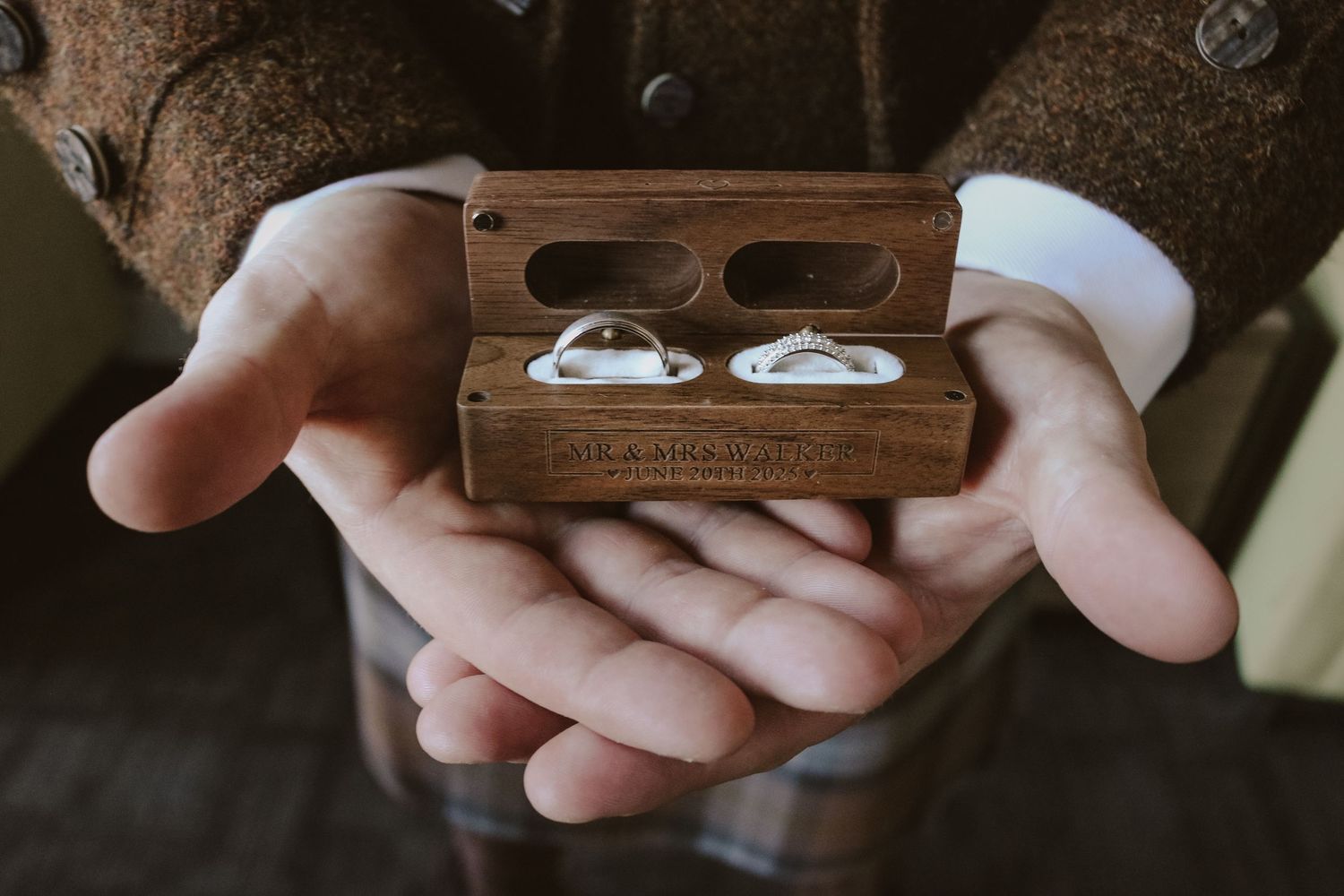 Hands gently holding a wooden box containing two wedding rings captured in moody lighting.