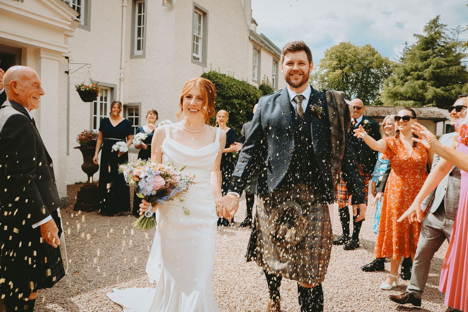 Wedding attendees celebrate by throwing white rice confetti outside a venue on a sunny day.