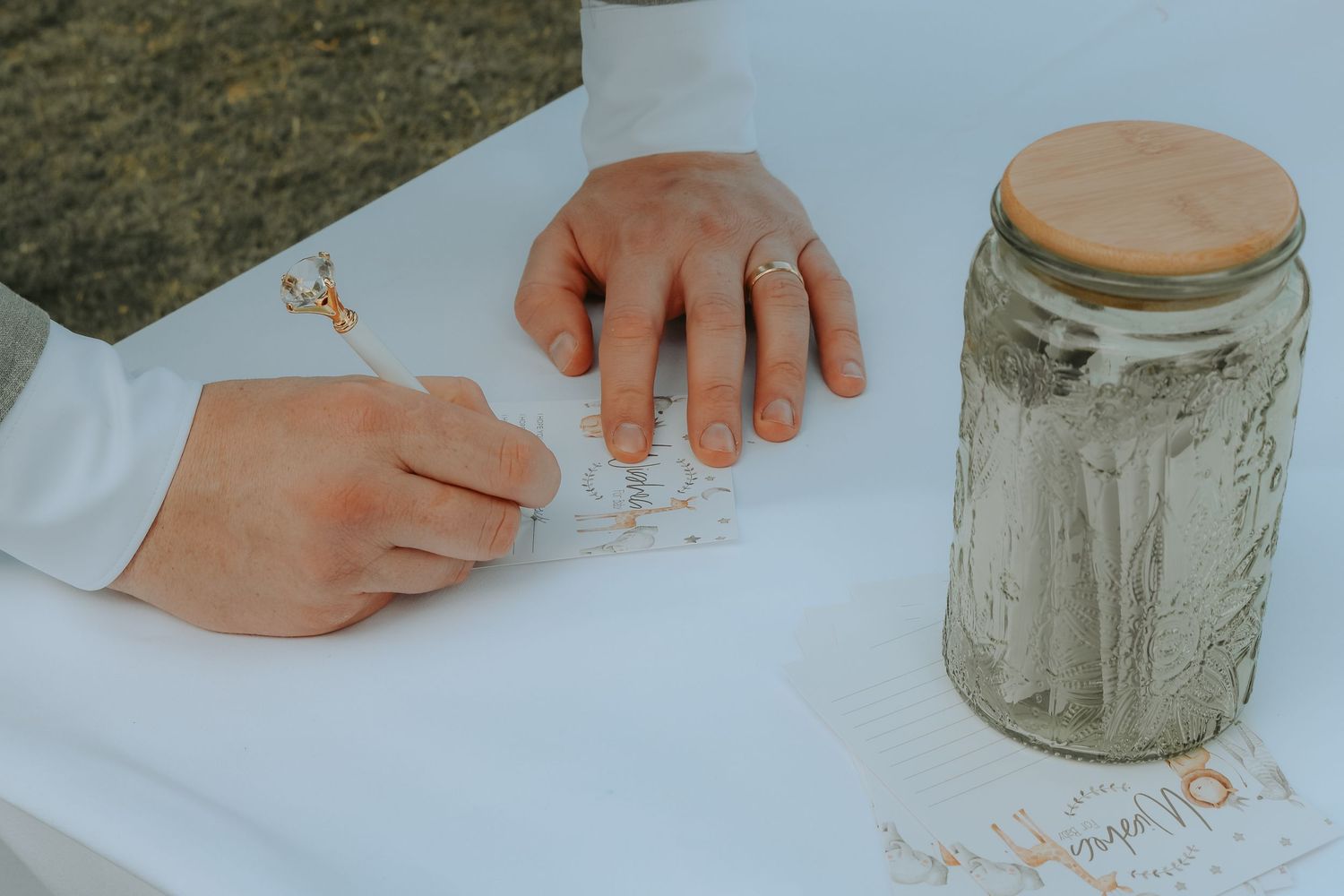 Hands writing on white paper next to a mason jar filled with grey material, one hand wearing a delicate ring.