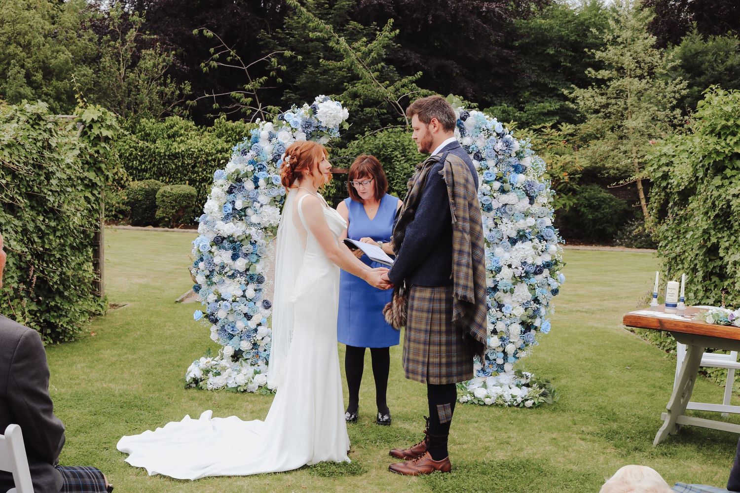 A wedding ceremony takes place outdoors in front of a floral arch decorated with soft blue and white flowers.