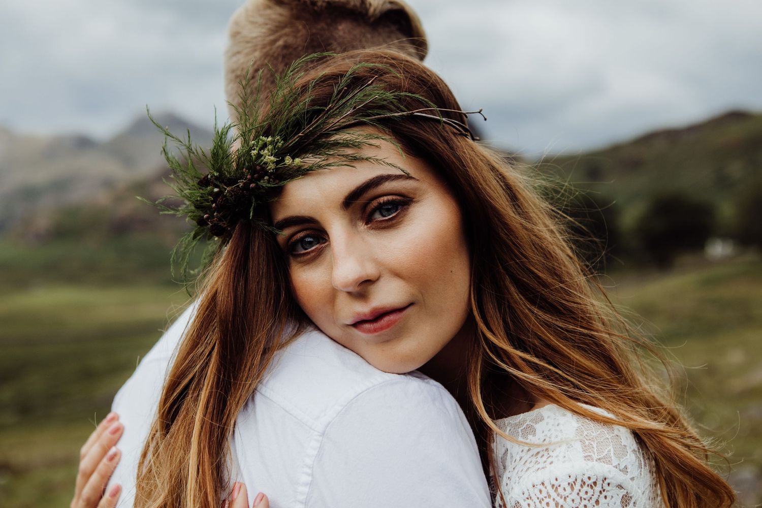 A woman in a white lace dress and leafy green crown poses against a scenic mountain landscape on a cloudy day.