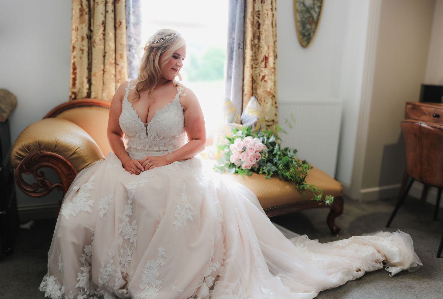 Elegant white lace wedding dress with deep V-neckline flows gracefully across vintage settee beside floral arrangement.