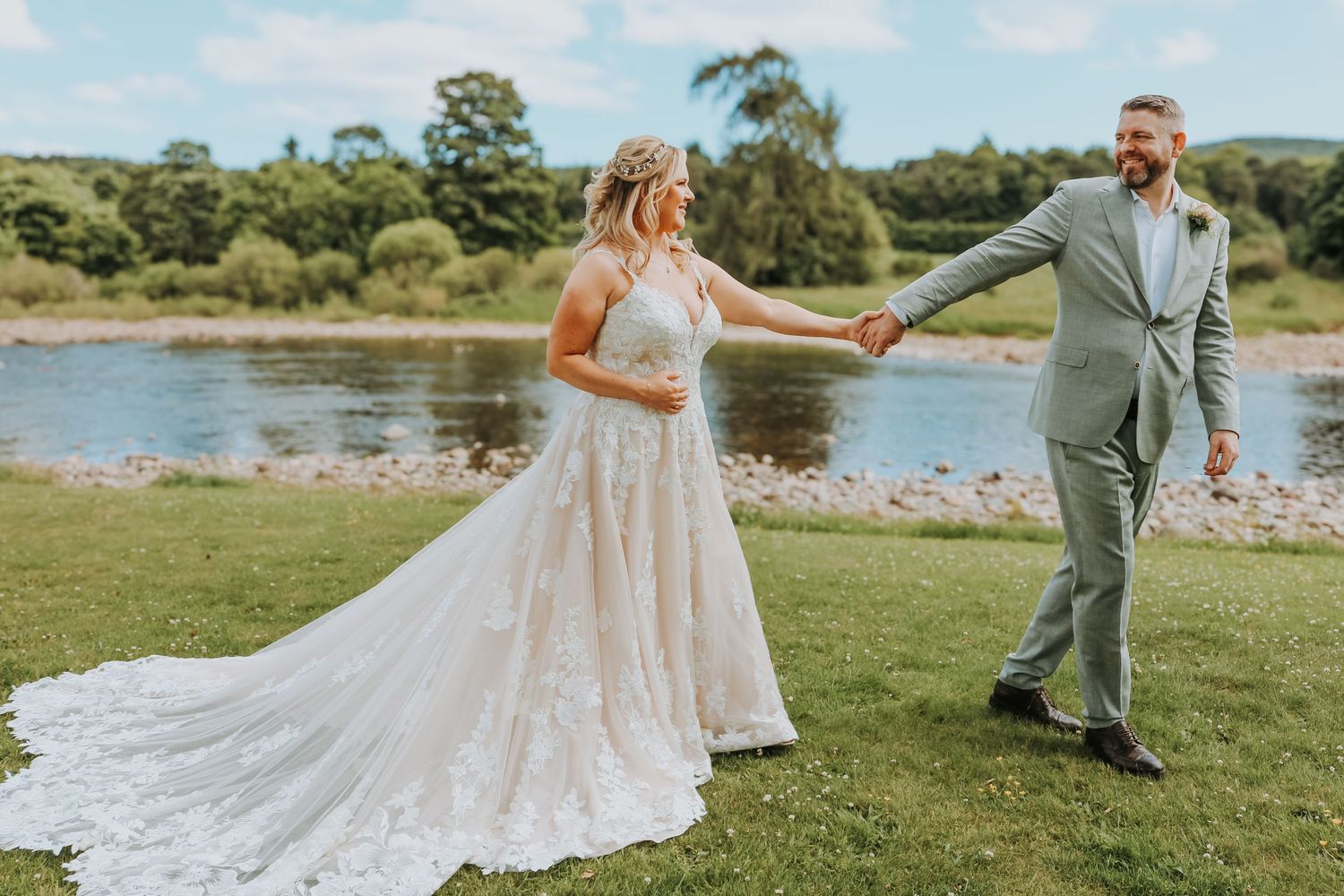 Joyful couple dancing together on grassy lawn by a scenic waterfront on their wedding day.