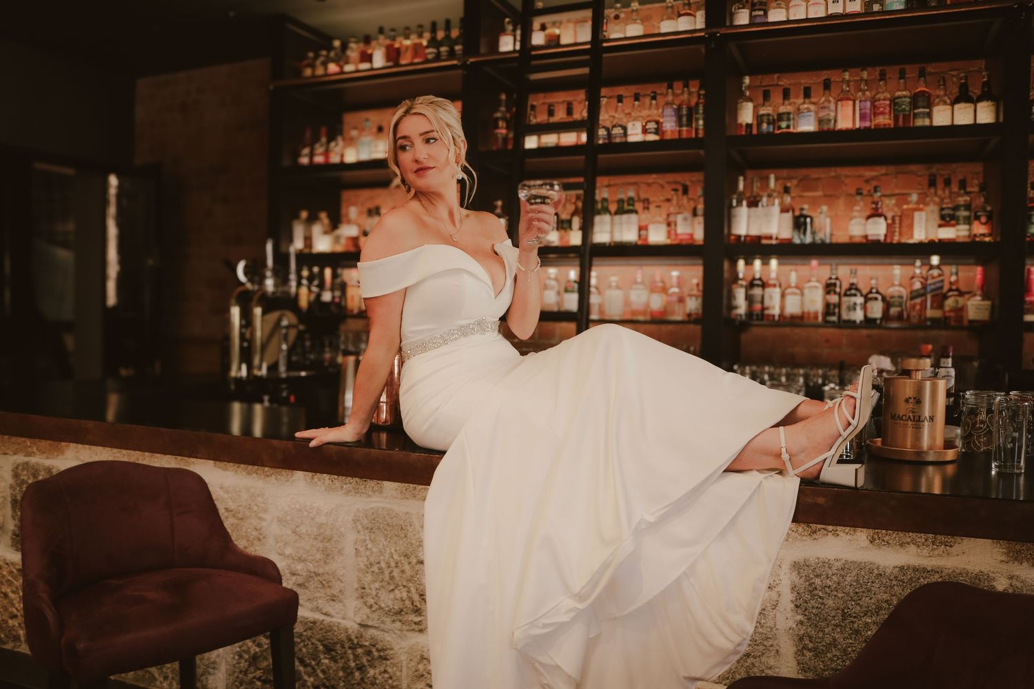 A bride in a white wedding dress sits elegantly on a bar stool in a rustic-style bar with wooden and stone decor.