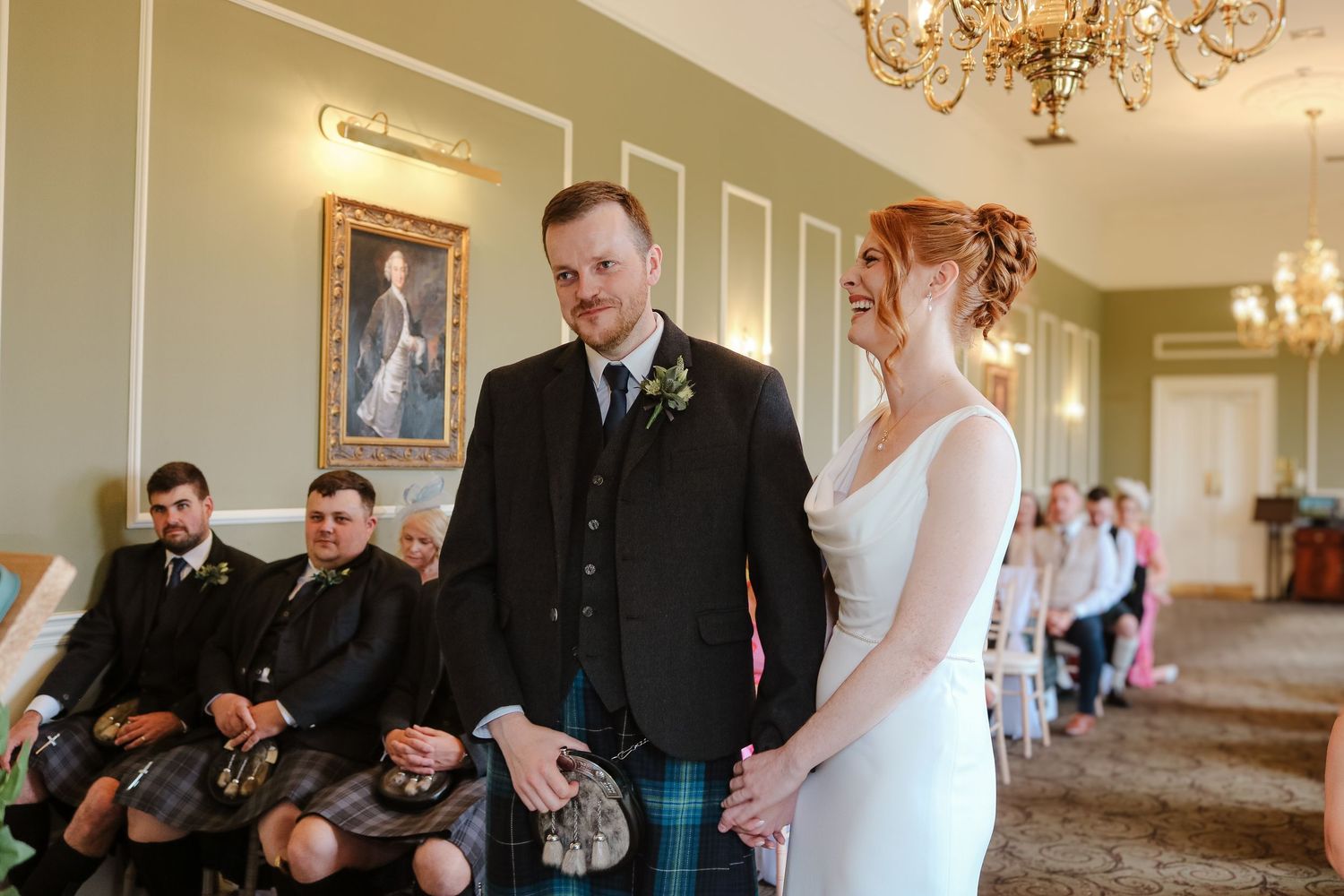 Wedding ceremony in an elegant room with traditional Scottish kilts and formal attire against pale green walls.
