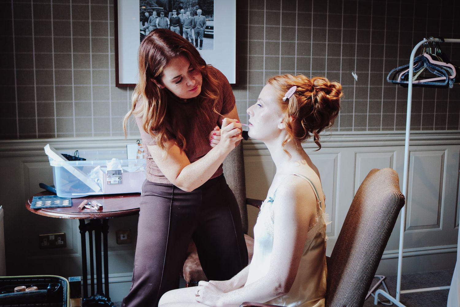 A makeup artist applies finishing touches to a bride's hair during wedding preparations in a vintage-style room.