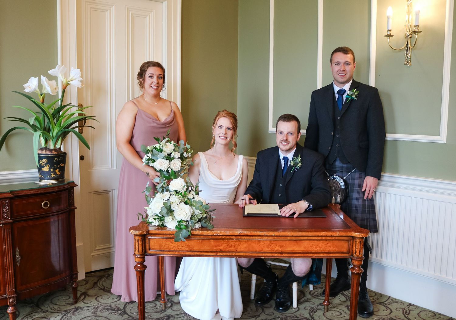 Wedding party gathered around an antique wooden desk during formal signing ceremony in an elegant room with sage green walls.