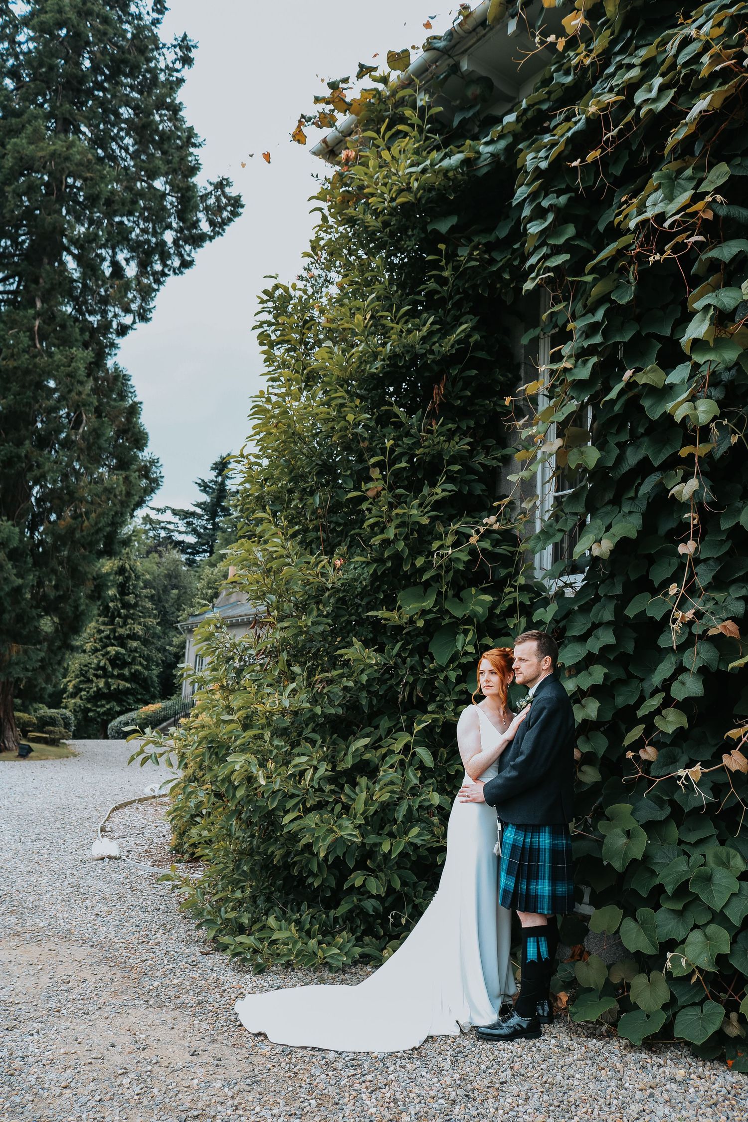 A romantic couple in wedding attire shares intimate moments on a gravel path bordered by lush green foliage.