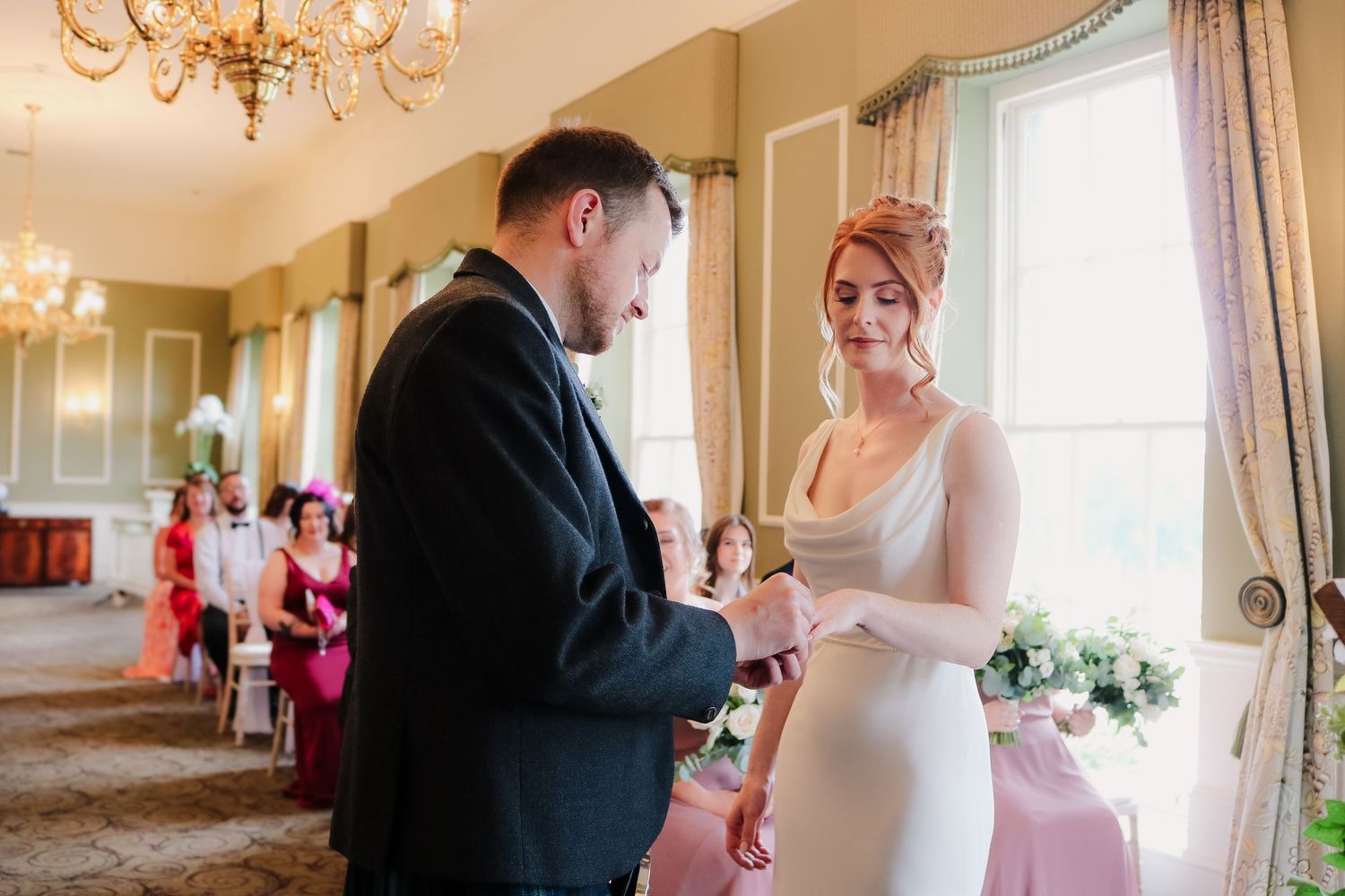 A couple exchanges rings during an elegant wedding ceremony in a bright room with chandelier and pink decor.
