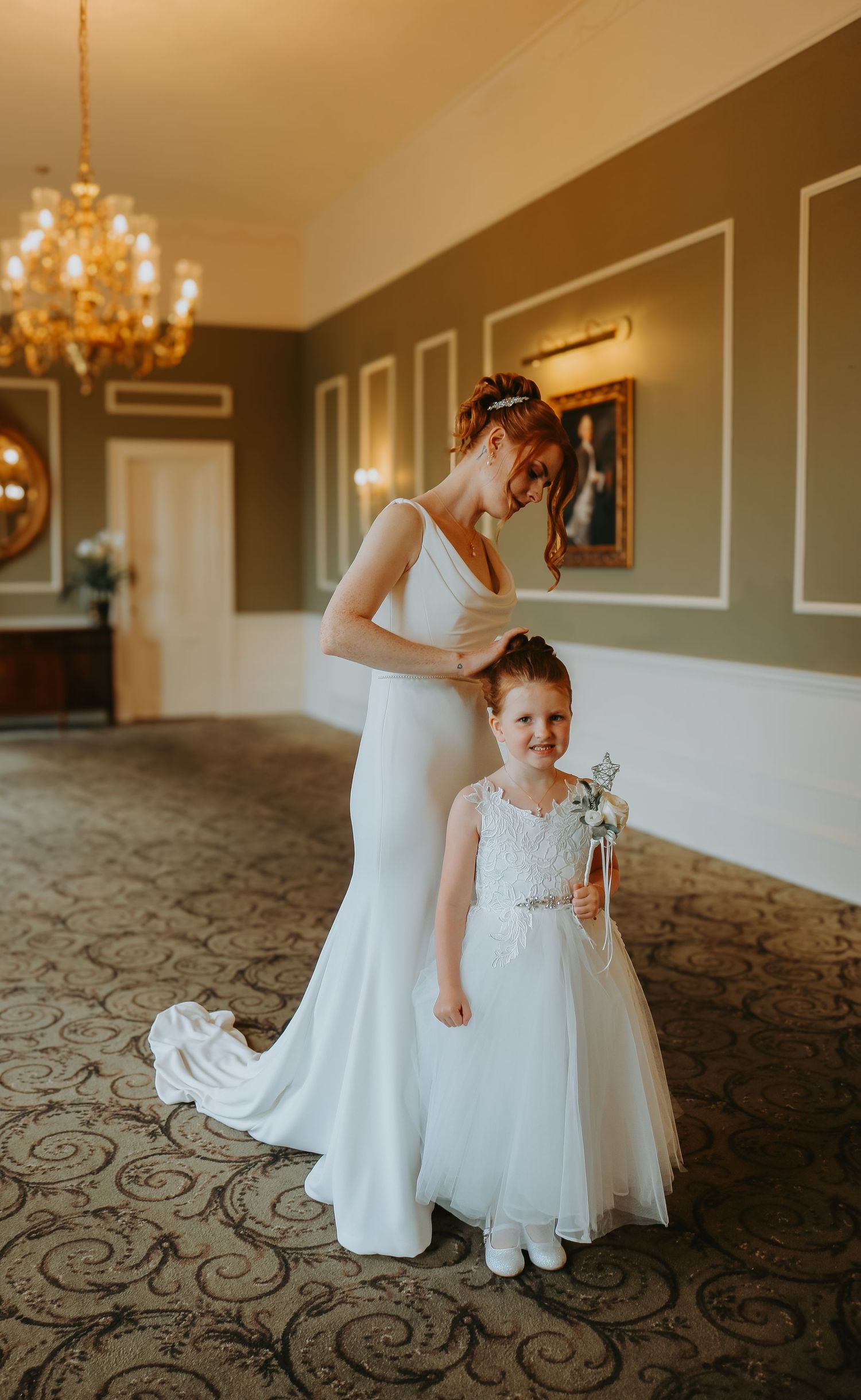 A bride and flower girl in matching white dresses share a sweet moment in an elegant hotel hallway with a chandelier.