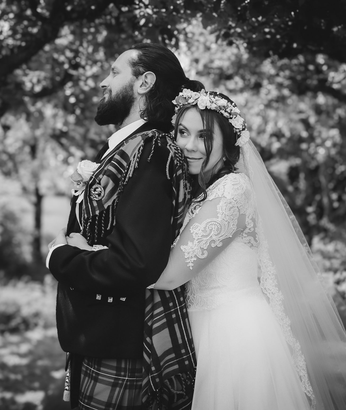 A black and white wedding photo showing a couple in traditional Scottish dress with kilt and lace gown embracing outdoors.