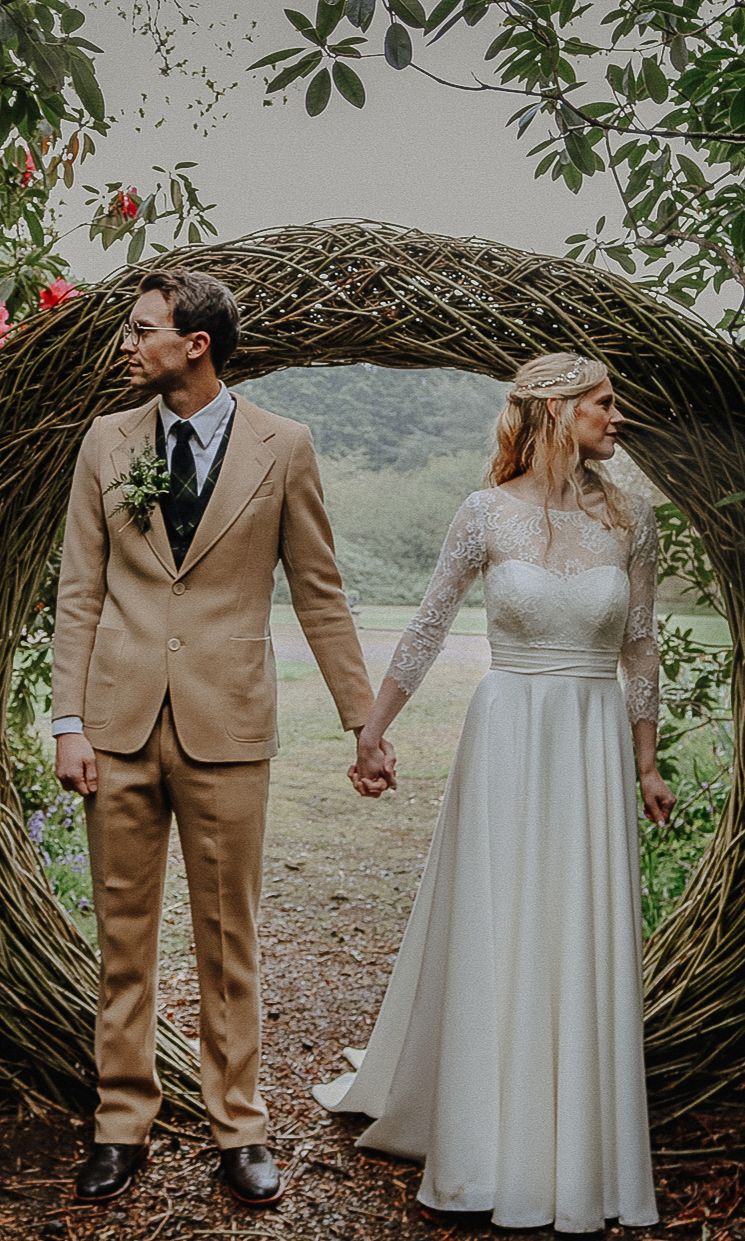 A newlywed couple holds hands beneath a rustic woven arch surrounded by greenery at an outdoor wedding ceremony.