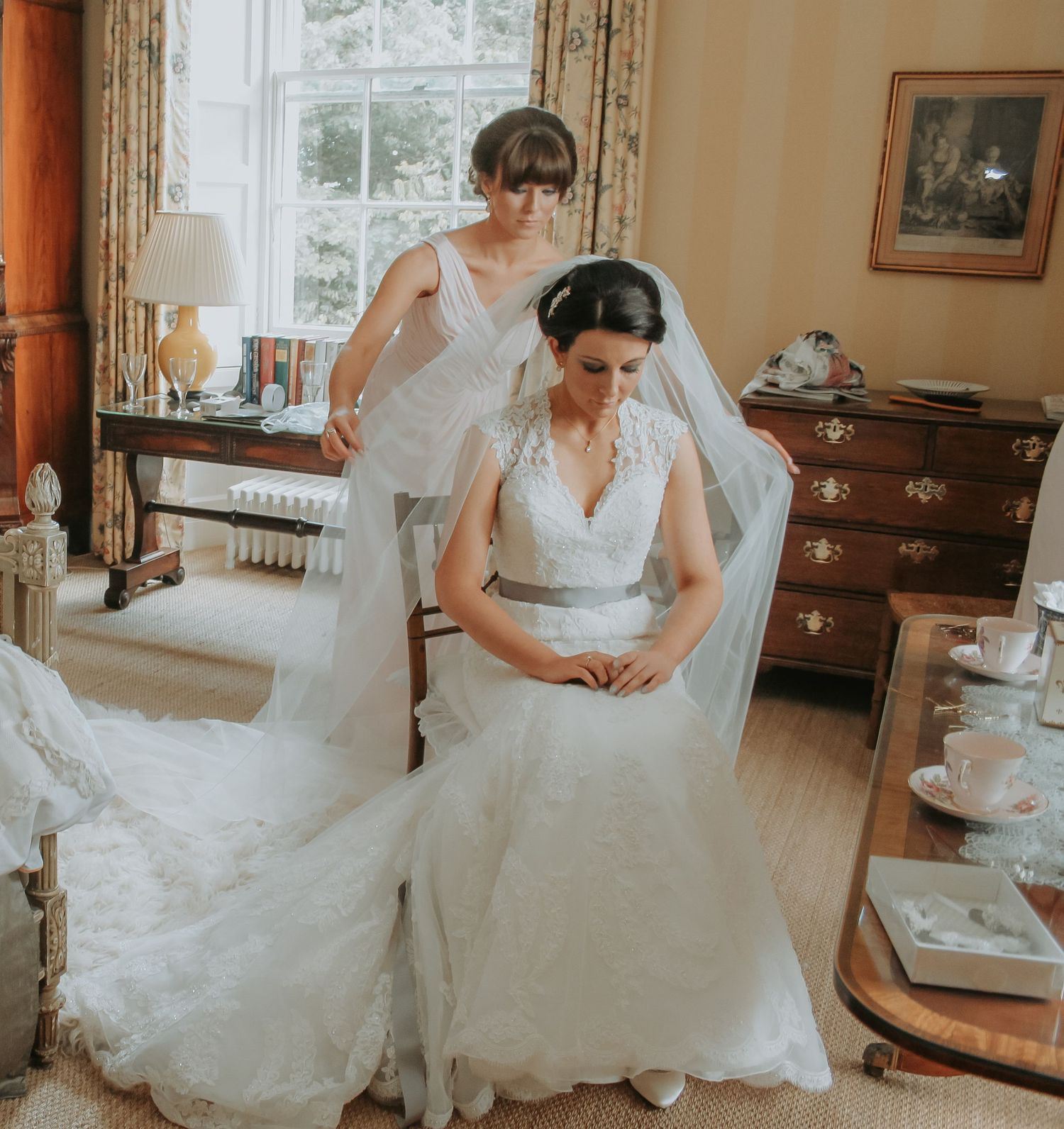 A bride in a lace white gown and veil sits in an elegant room with floral curtains while getting ready for the ceremony.