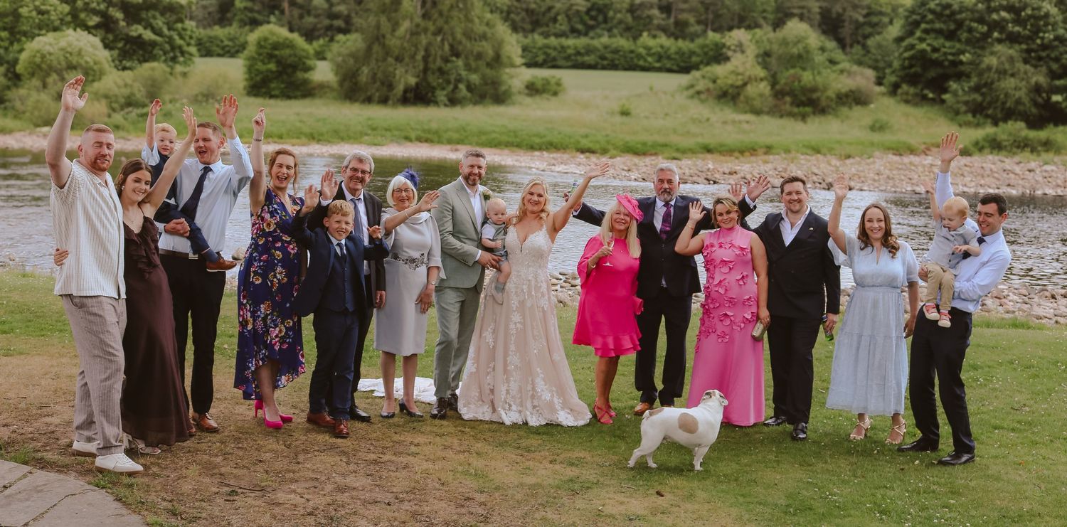Happy wedding party in formal attire poses together outdoors by a river on a sunny summer day.