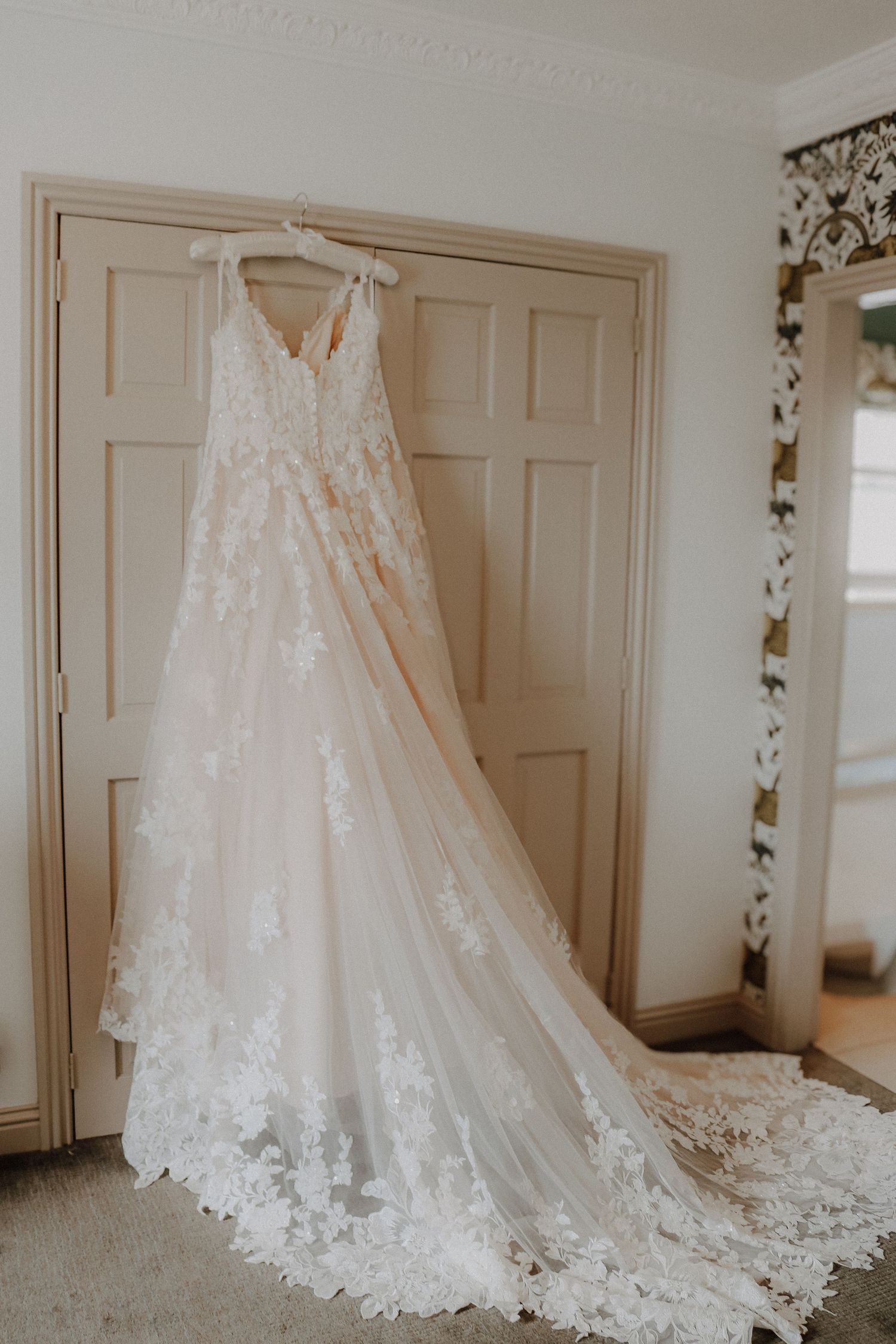 Wedding dress with intricate lace details and long train hangs elegantly against white paneled door in natural light.