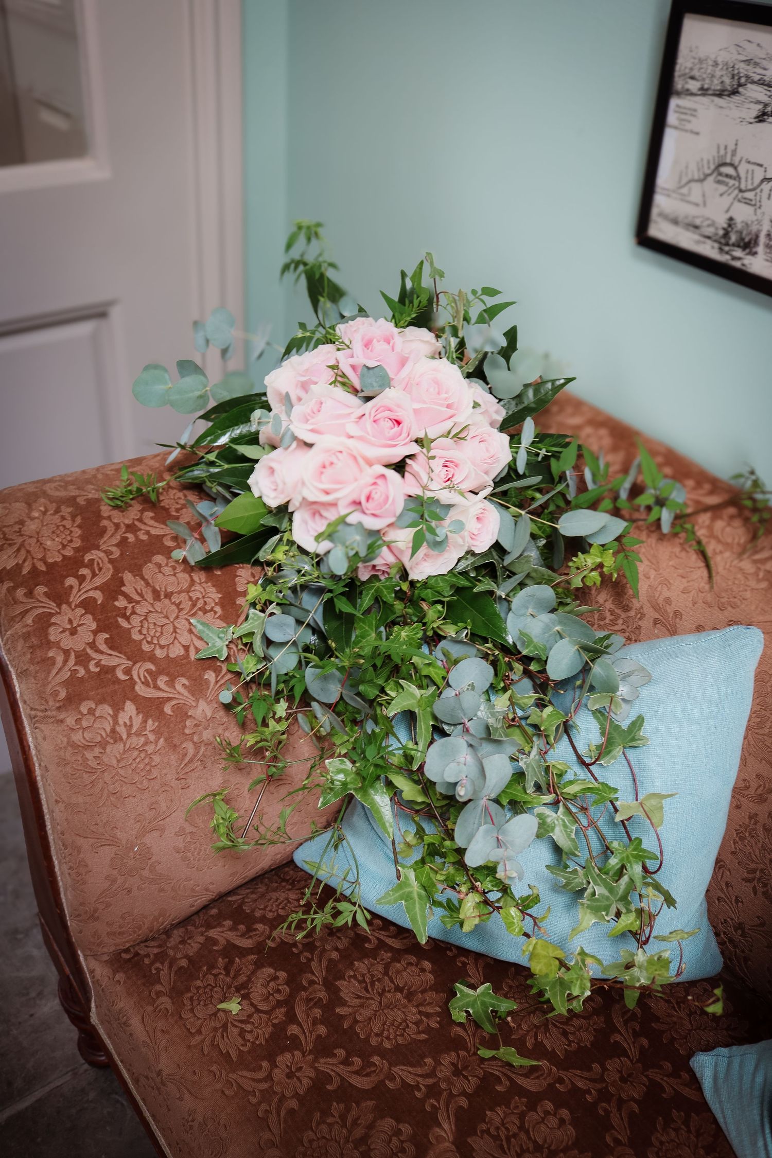 Pink roses and eucalyptus bouquet arranged with trailing greenery rests on a brown damask upholstered surface.