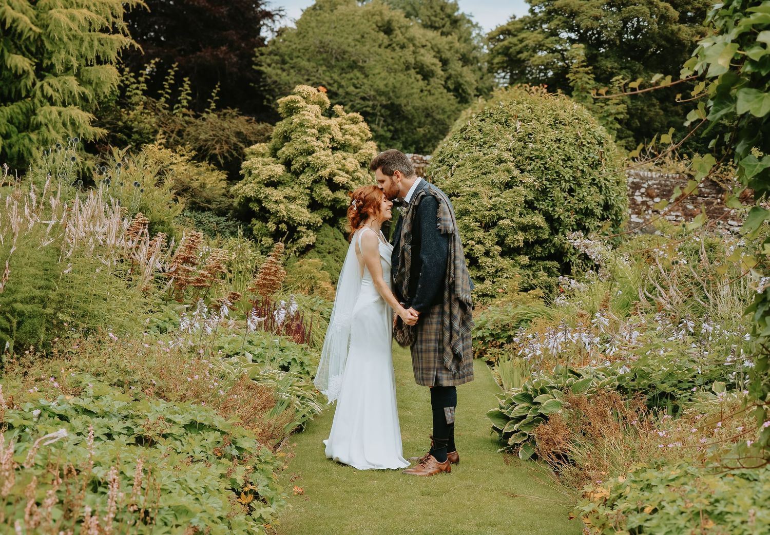 A romantic garden wedding scene with a figure in traditional Scottish kilt and a bride in a flowing white gown among lush foliage.