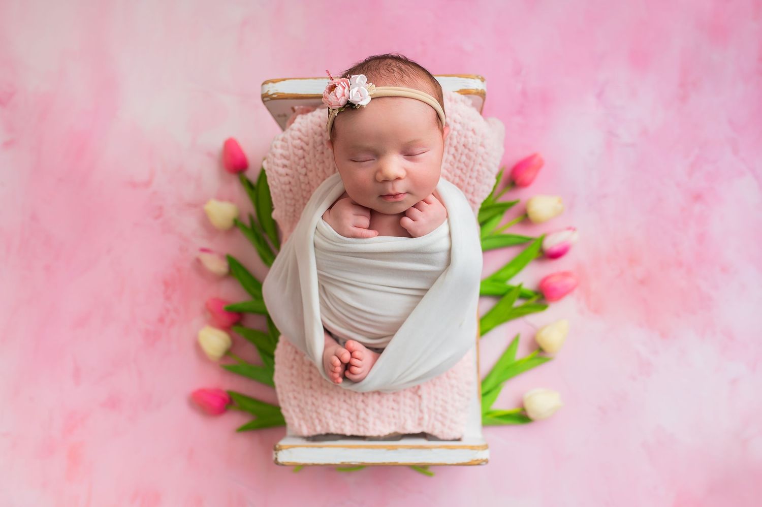 Newborn baby wrapped in white swaddle lying on pink blanket with tulips in a soft pink studio setting.