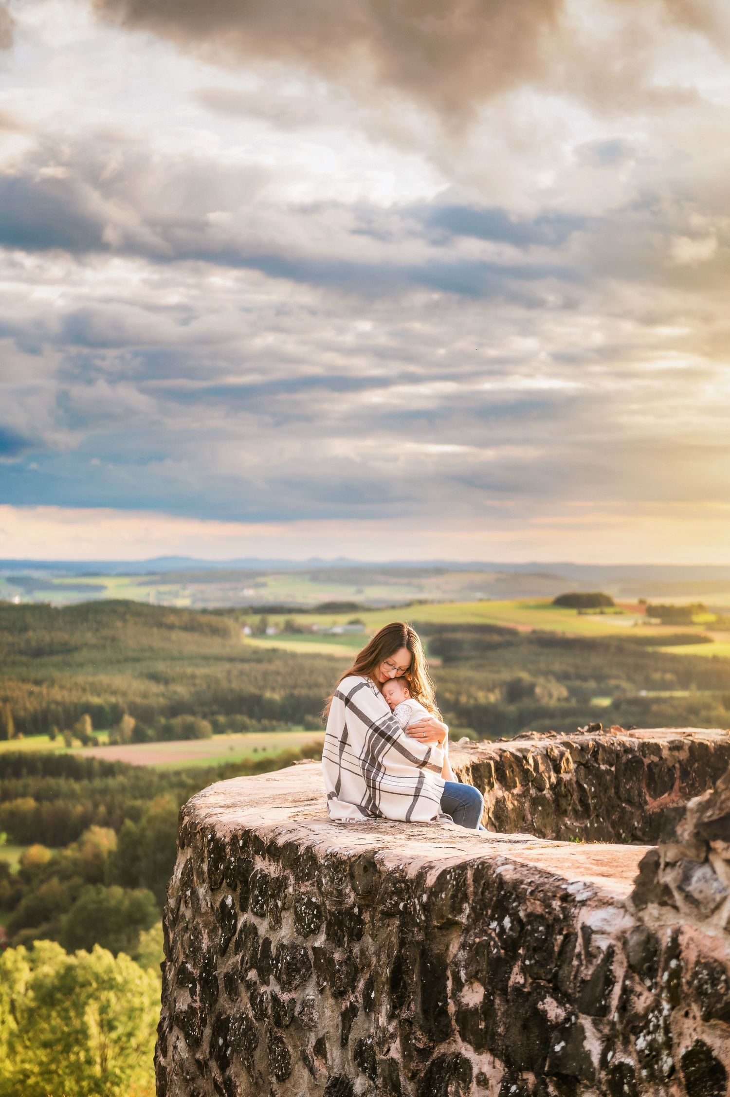 A figure wrapped in a blanket sits on a rocky cliff edge overlooking a vast landscape at sunset.