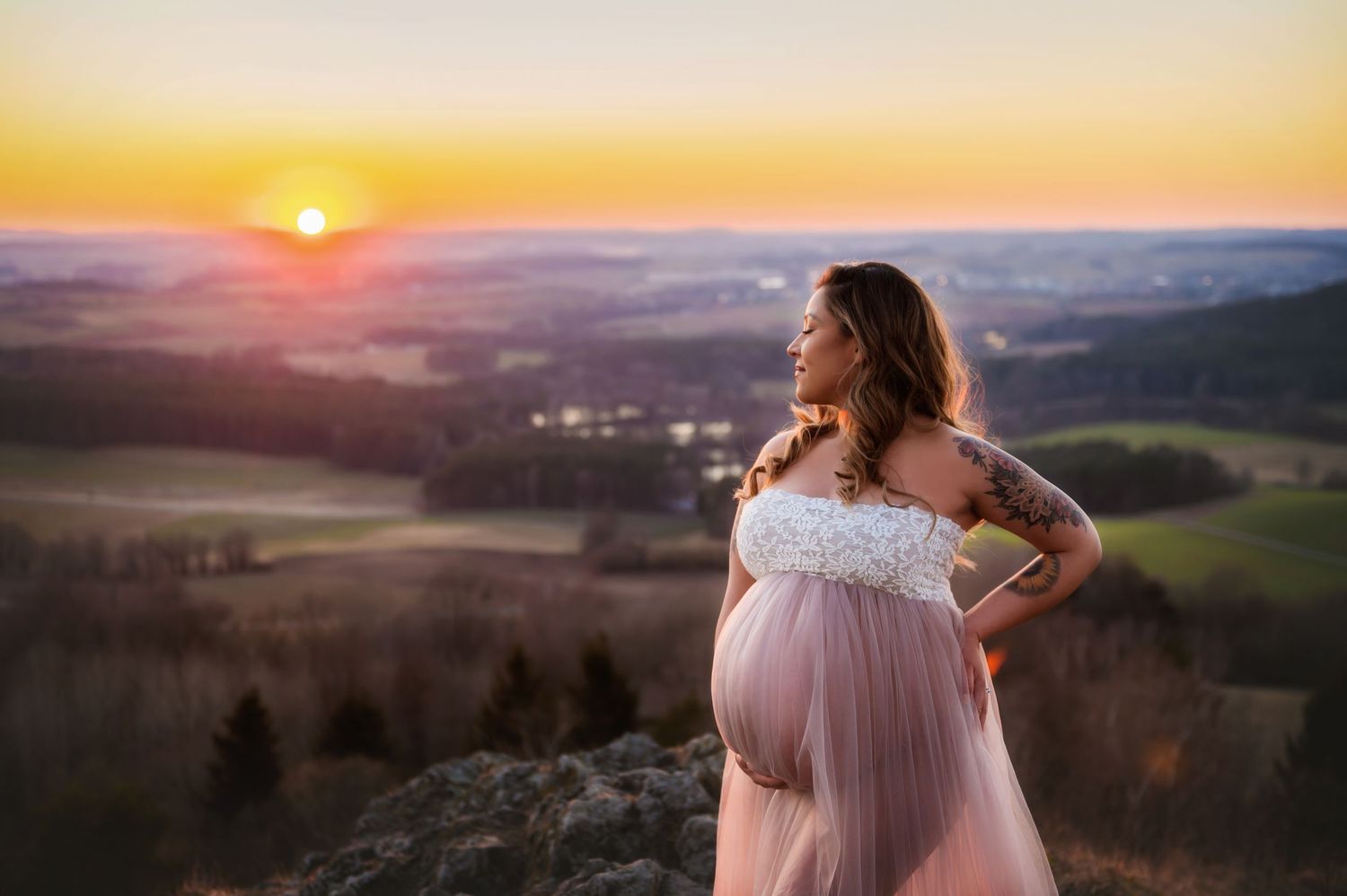 A silhouetted person in a flowing pink dress stands on a mountain overlook during a golden sunset over misty rolling hills.