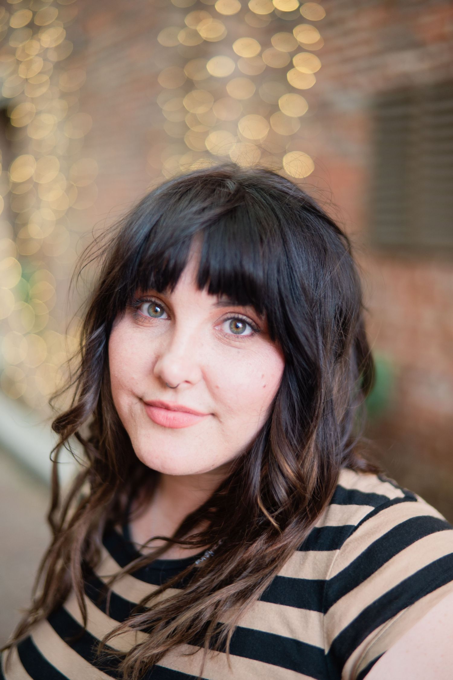 A person with dark wavy hair and bangs smiles softly while wearing a black and tan striped shirt against a bokeh background.