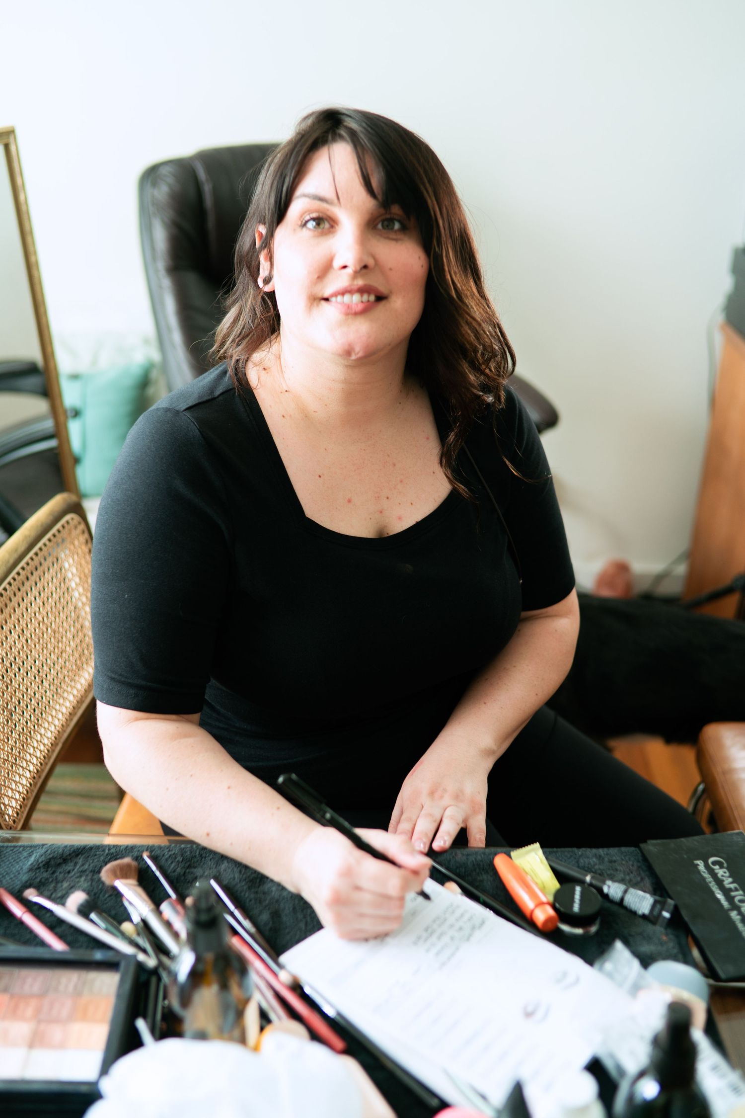 A woman in a black shirt sits at a desk with makeup and cosmetic tools while writing on paper.