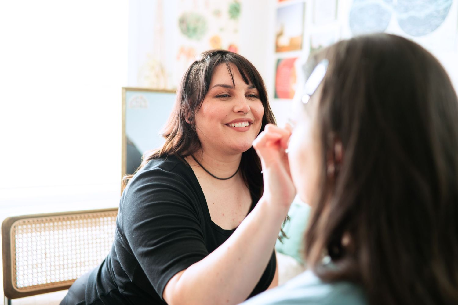 A makeup artist in a black sweater applies cosmetics to a client in a bright indoor setting.