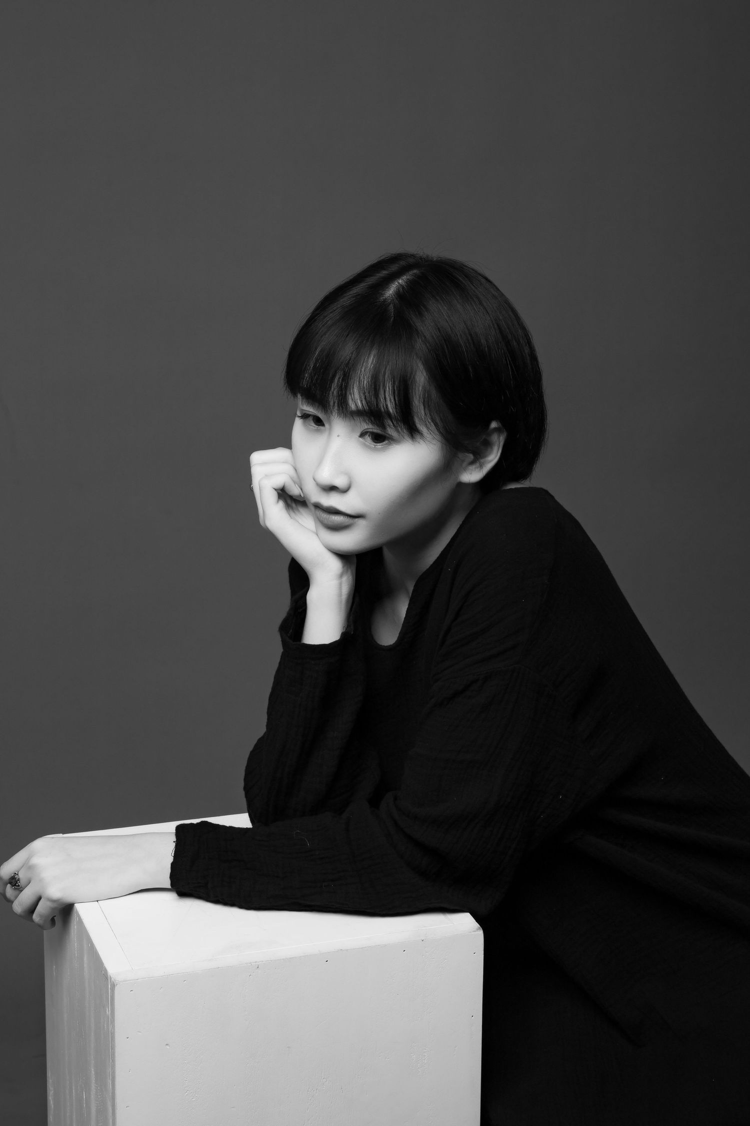 Black and white portrait of someone in dark sweater leaning on white cube with contemplative pose against gray backdrop.